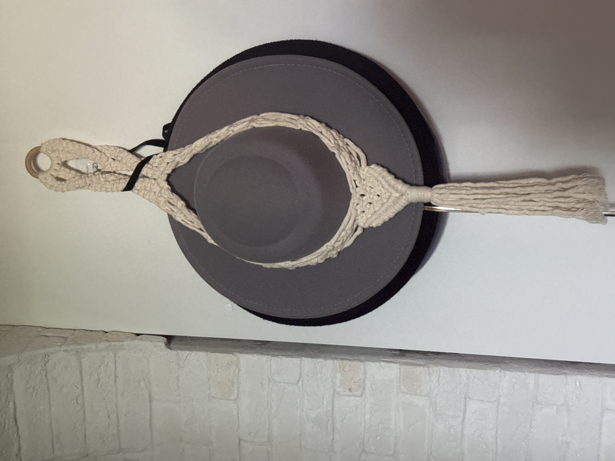 A gray and black hat hangs on a white wall, supported by a macrame hanger. The hat is a wide-brimmed fedora style, with a gray body and a black brim edge. The macrame hanger is a light beige color, featuring a wooden ring at the top for hanging and a tassel at the bottom. The wall behind the hat is white, and a brick wall section is visible below. No specific location in Kazan, Russia, or any people, subjects, actions, or interactions are evident in the image.
