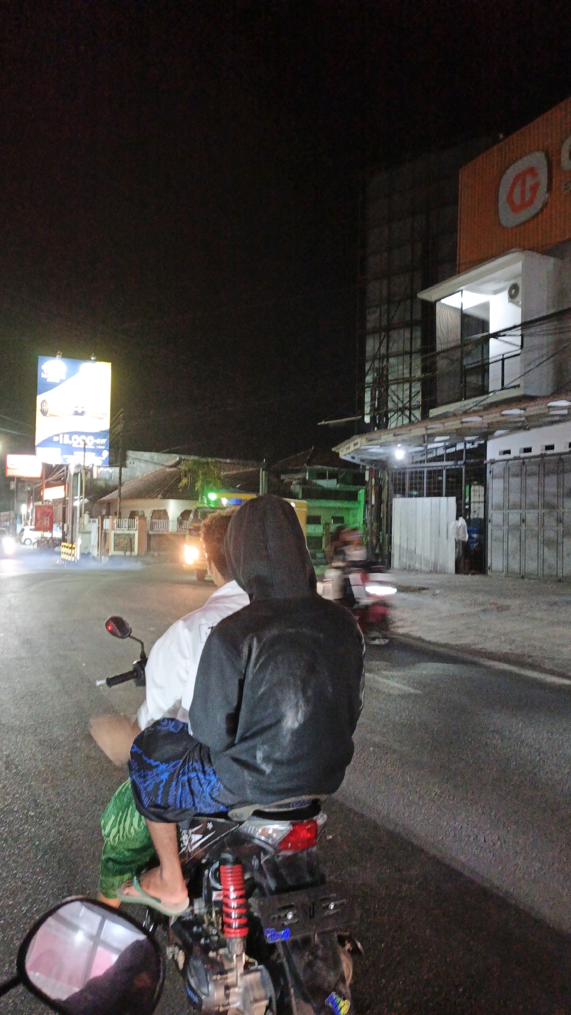This is a nighttime, low-light, vertical, first-person perspective shot of two individuals on a motorcycle in Desa Karangbenda, Indonesia. The motorcycle is facing away from the camera and is on an asphalt road. The rider in the back is wearing a black hoodie and blue patterned shorts, while the rider in front is wearing a white long-sleeved shirt and green pants. The rear of the motorcycle, including its red shock absorber and taillight, is visible. In the background, there is a modern building with orange and white signage. To the left, a large illuminated billboard advertising a product or service stands out. The road is lined with buildings and other motorcycles, some of which are in motion, indicated by motion blur. The scene appears to be in an urban or semi-urban setting at night, illuminated by artificial lights.