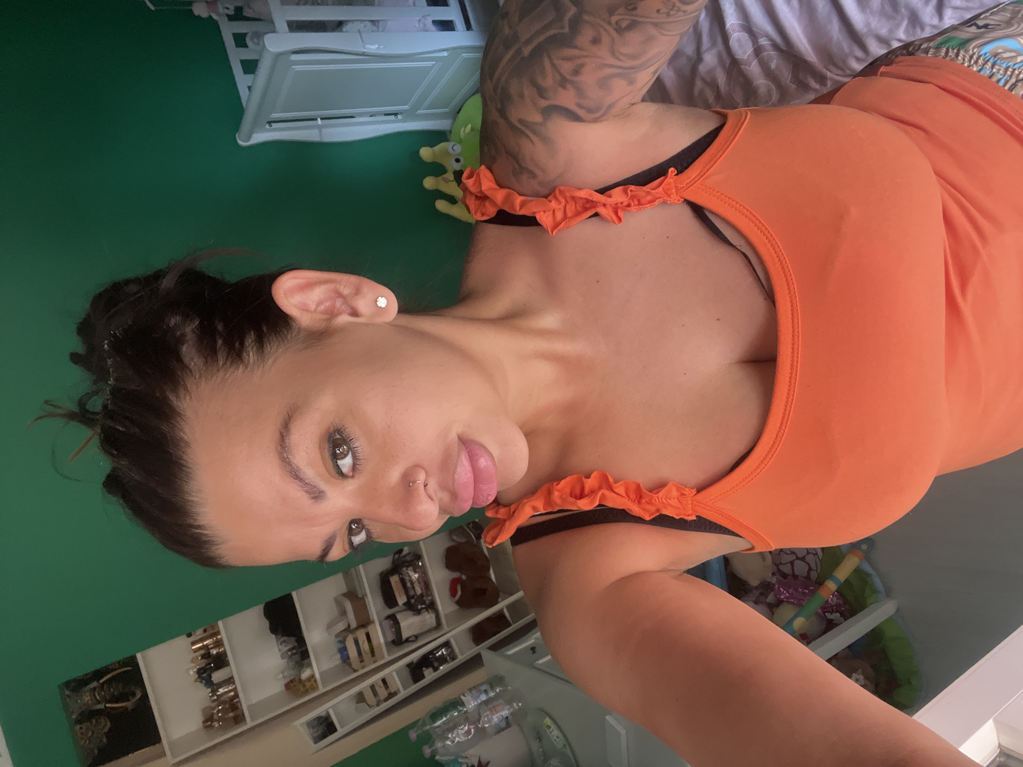 A woman with dark hair pulled up in a bun and a nose ring is looking at the camera. She is wearing an orange tank top with ruffled straps. Her left arm has a large tattoo. In the background, there is a white crib, a bookshelf filled with various items, and a green wall. To the left, a dresser with bottles of water is visible. The scene appears to be indoors, possibly in a bedroom.