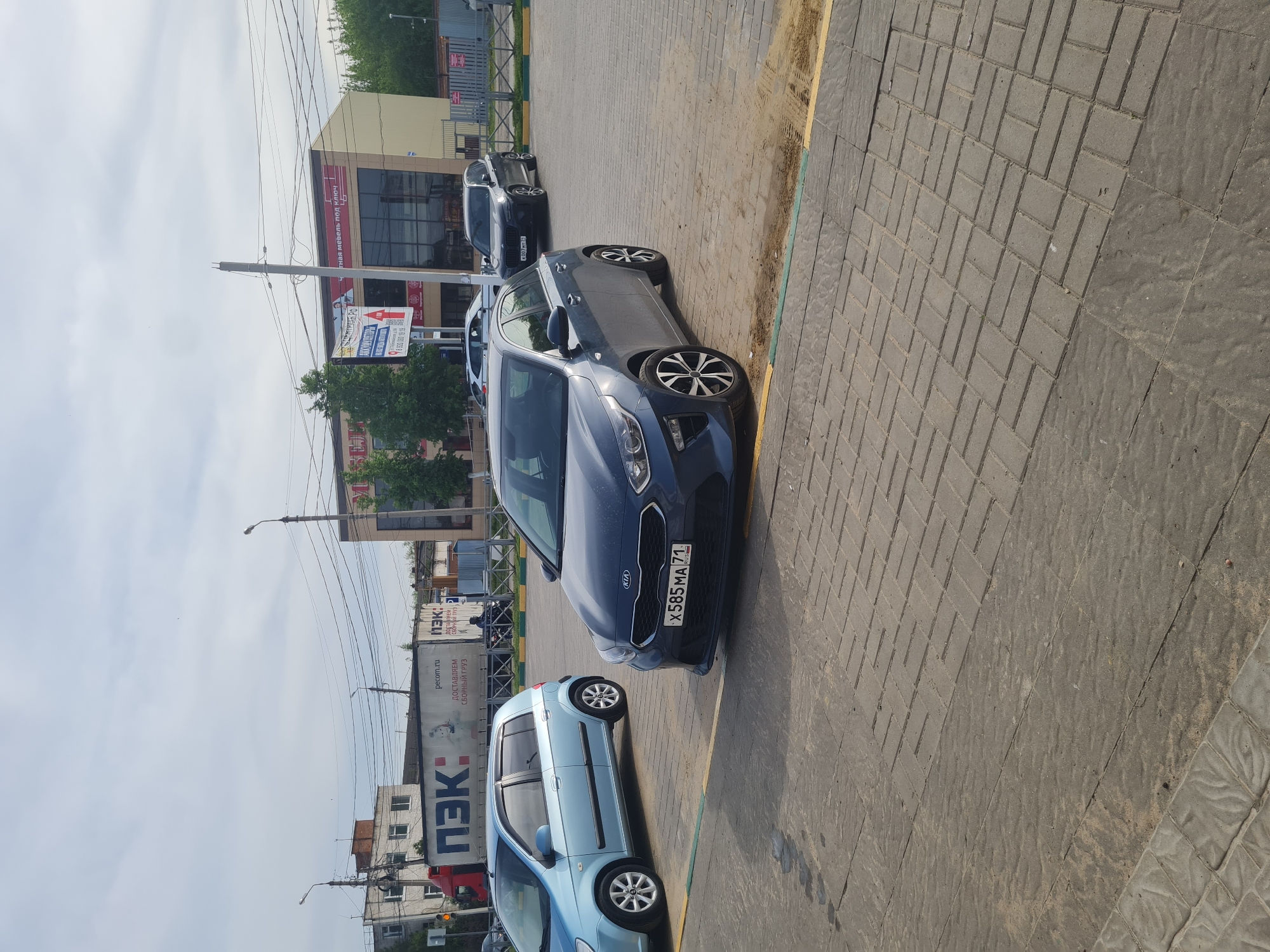 This media file captures a daytime street scene in Novomoskovsk, Russia, under an overcast sky. The image is taken from a slightly elevated perspective, looking down a paved area adjacent to a road.
In the foreground, a dark blue Kia hatchback is prominently parked on a paved surface composed of interlocking grey concrete blocks, with a yellow line marking the edge. The car's Russian license plate, X585 MA 71, is clearly visible. Behind it, a light blue hatchback is also parked. Further along the street, a dark grey SUV is visible.
In the mid-ground, commercial buildings line the street. One building features a large sign with the partially visible Cyrillic text "тная мебель под ключ," indicating a furniture business. Another advertising banner with numbers like "12:4200" and "000 89 19" is also present. Utility poles and a network of power lines crisscross the sky above the street.
The background on the left features a large white truck from the Russian logistics company "ПЭК" (PEK). The truck's side displays the company logo and the text "pecom.ru" and "ДОСТАВЛЯЕМ СБОРНЫЙ ГРУЗ" (Delivering Groupage Cargo). The sky is bright and cloudy, characteristic of an overcast day. The overall scene depicts a typical commercial or urban environment.