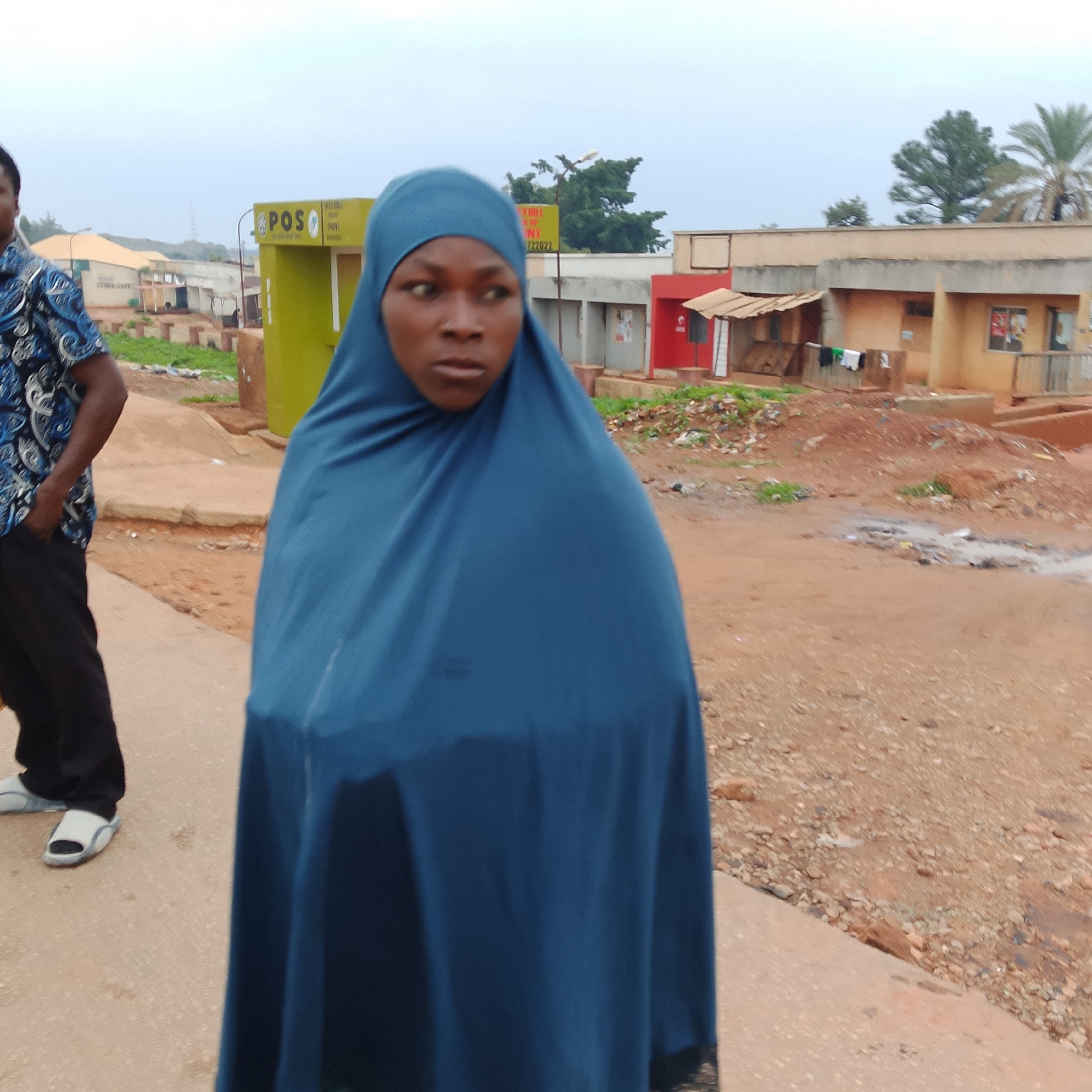 An outdoor, eye-level medium shot captures a person wearing a blue hijab standing slightly to the right of the frame, facing forward with their head turned to the right. Their left arm is bent, and their hands are not visible. To the left of the frame, a person wearing a blue patterned shirt and black pants stands with their left hand in their pocket, also facing forward. The background features a street with a dirt shoulder, a light-colored sidewalk, and buildings with signs. One sign reads "POSO" in yellow letters on a green kiosk. A second sign above it has yellow text on a green background. Another building to the right is a light brown structure with a red section on the left. Trees, including palm trees, are visible in the distance. The sky is overcast and light. The ground is uneven, with some puddles and debris.