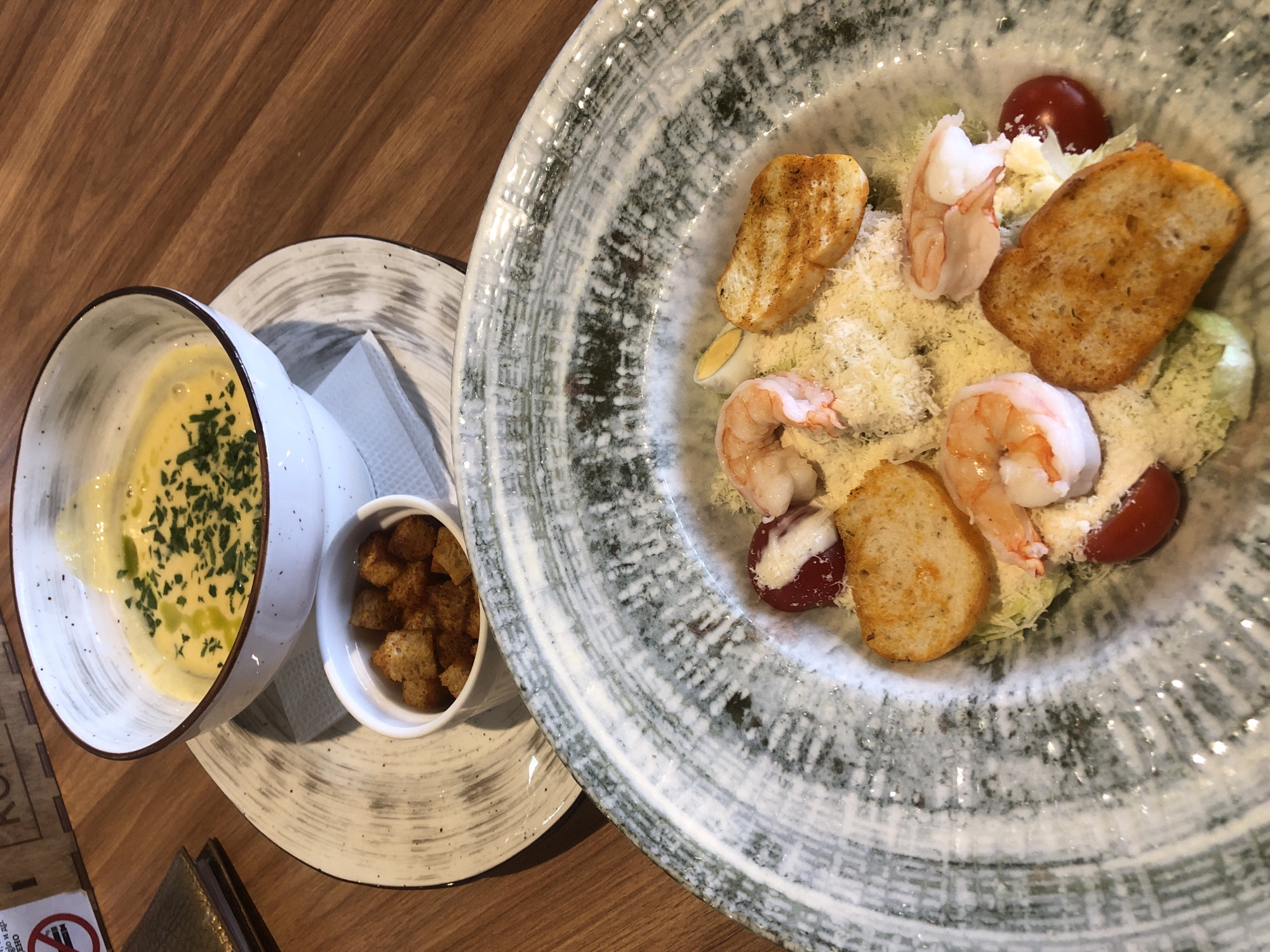 A close-up, overhead shot captures a culinary scene on a wooden table in Izhevsk, Russia. In the foreground, a large, rustic ceramic plate holds a Caesar salad featuring several cooked shrimp, cherry tomatoes, croutons, shredded cheese, and lettuce. In the background, a white bowl contains a creamy yellow soup garnished with chopped herbs and a drizzle of oil. A small white dish of croutons sits beside the soup bowl, which rests on a patterned charger plate. The overall impression is of a neatly presented, hearty meal.