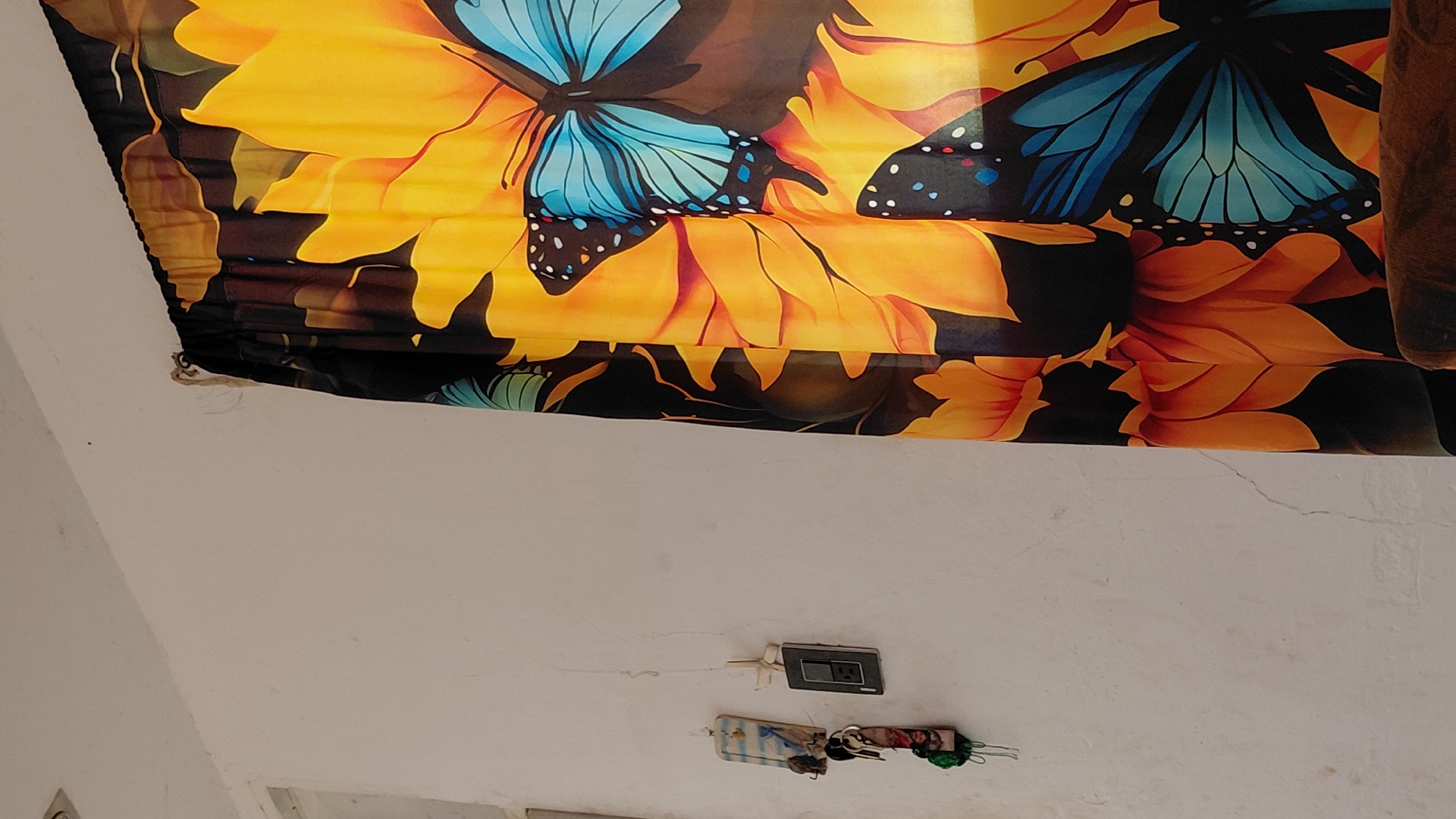 An indoor scene shows a white wall with a dark switch plate and a set of keys hanging from a hook on the left. To the right, a curtain with a vibrant sunflower and butterfly print is partially visible. The curtain is adorned with large yellow sunflowers and multiple blue butterflies with black spots. The scene is brightly lit.