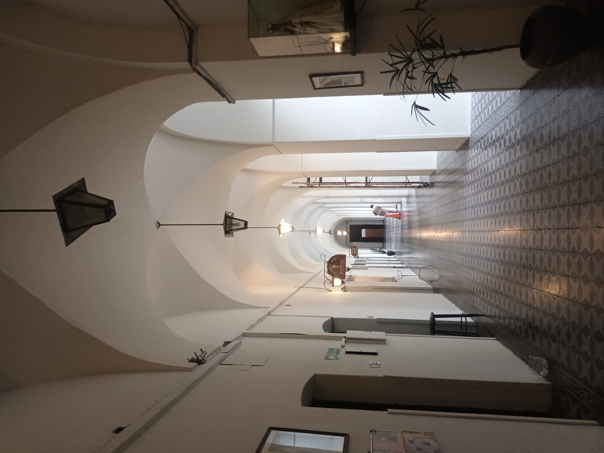 A long, wide hallway with arched ceilings is the primary focus of this indoor shot. The hallway appears to stretch into the distance, diminishing in size due to perspective. The ceilings are painted white, and evenly spaced light fixtures hang down the length of the corridor. The walls are also white, and adorned with a mix of framed pictures, display cases, and signage. On the right side of the hallway, a patterned tile floor adds visual detail to the scene.
At the far end of the hallway, a person is visible, sitting on a red stool. The person's actions or specific features are hard to discern due to the distance. The environment is bright, with the hallway illuminated by the light fixtures and natural light coming from the far end of the passage.
Based on visual clues, particularly the architecture, signage, and décor, the location is likely a historic building or institution in Buenos Aires, Argentina. Specific identification requires further information, but the style is consistent with architectural details found in various parts of the city.