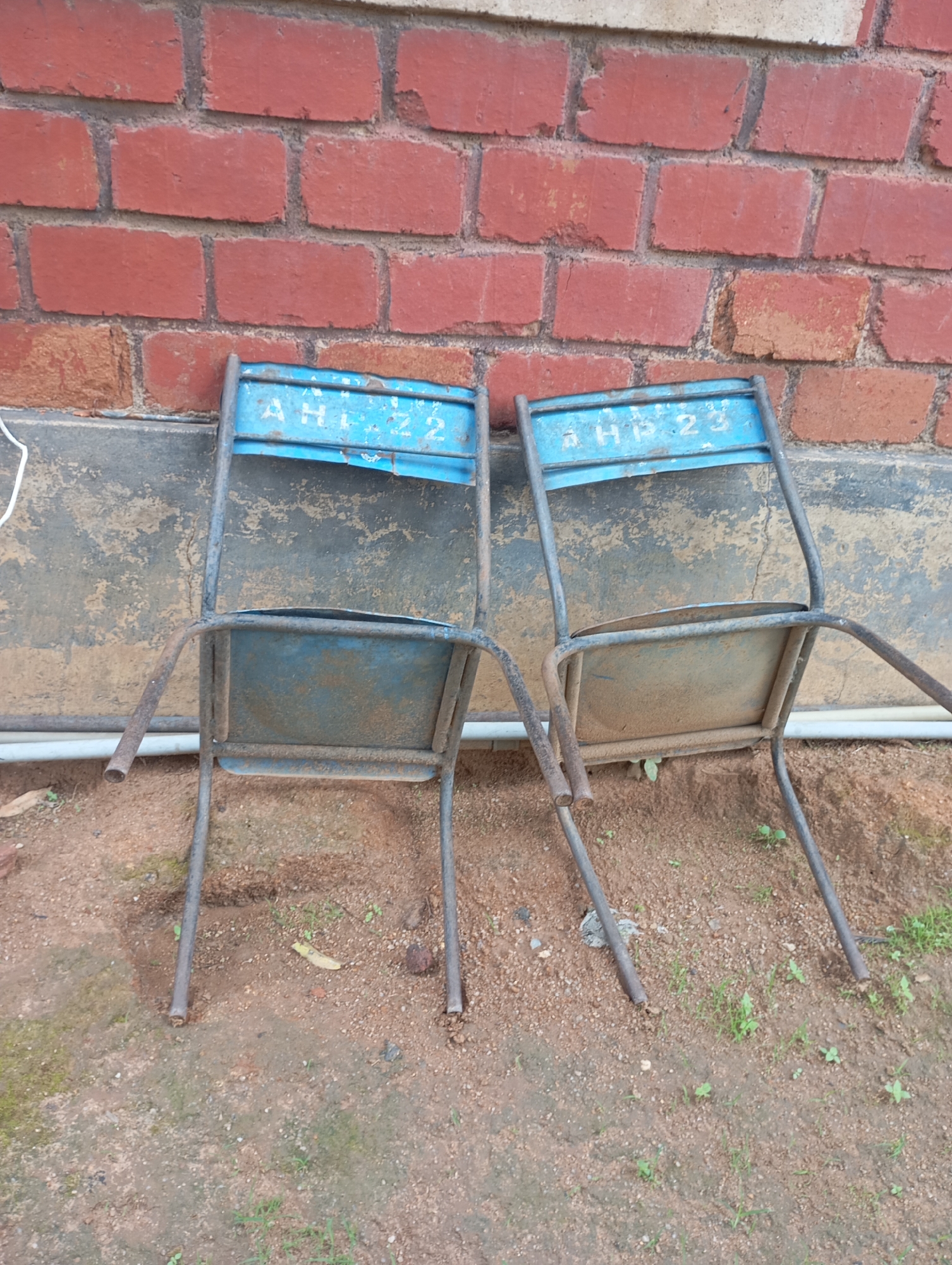 Two worn, blue metal chairs are positioned against a weathered brick wall in an outdoor setting. The chairs appear old and have visible signs of rust and dirt. The left chair has the markings "AHL 72" stenciled on its backrest, while the right chair has "AHL 23." The wall is constructed of red bricks with gray mortar. Below the chairs, the ground is a mixture of dirt and gravel with sparse green vegetation. The scene suggests an outdoor area, possibly a schoolyard or a community space, in New Stadium, Nigeria.