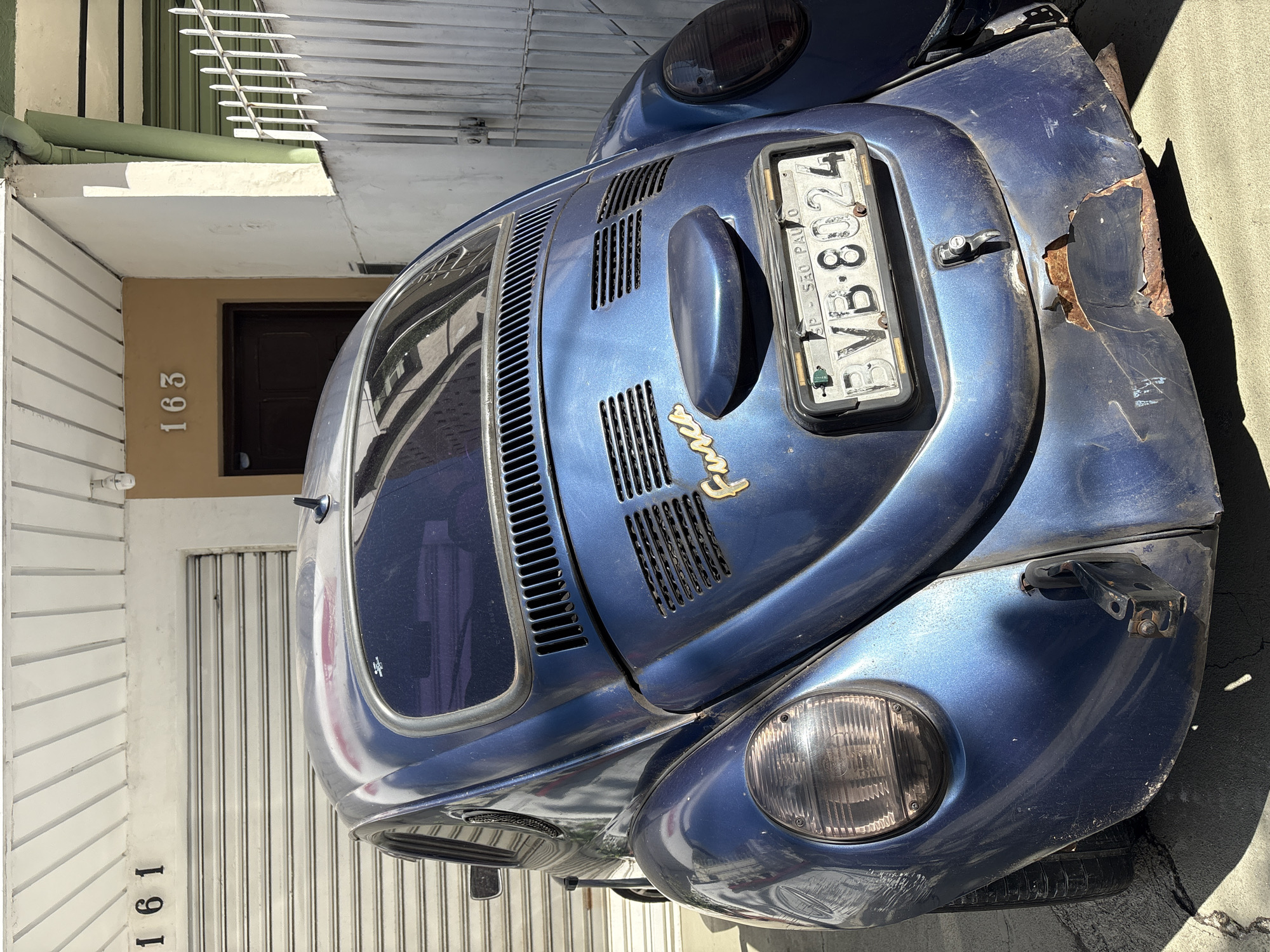 The image shows a rear view of a blue Volkswagen Beetle parked on a cracked concrete surface in front of a building. The building has white and orange sections. The numbers "161" are visible on the left side of the building, above a rolled-down garage door. The numbers "163" are visible on the right side, above a dark brown door. To the right of the door is a white metal fence. The car's license plate reads "BVB-8024 SP SAO PAULO". The car shows signs of rust and damage on the rear bumper. The overall scene suggests an urban environment in São Paulo, Brazil. There are no people visible in the image.