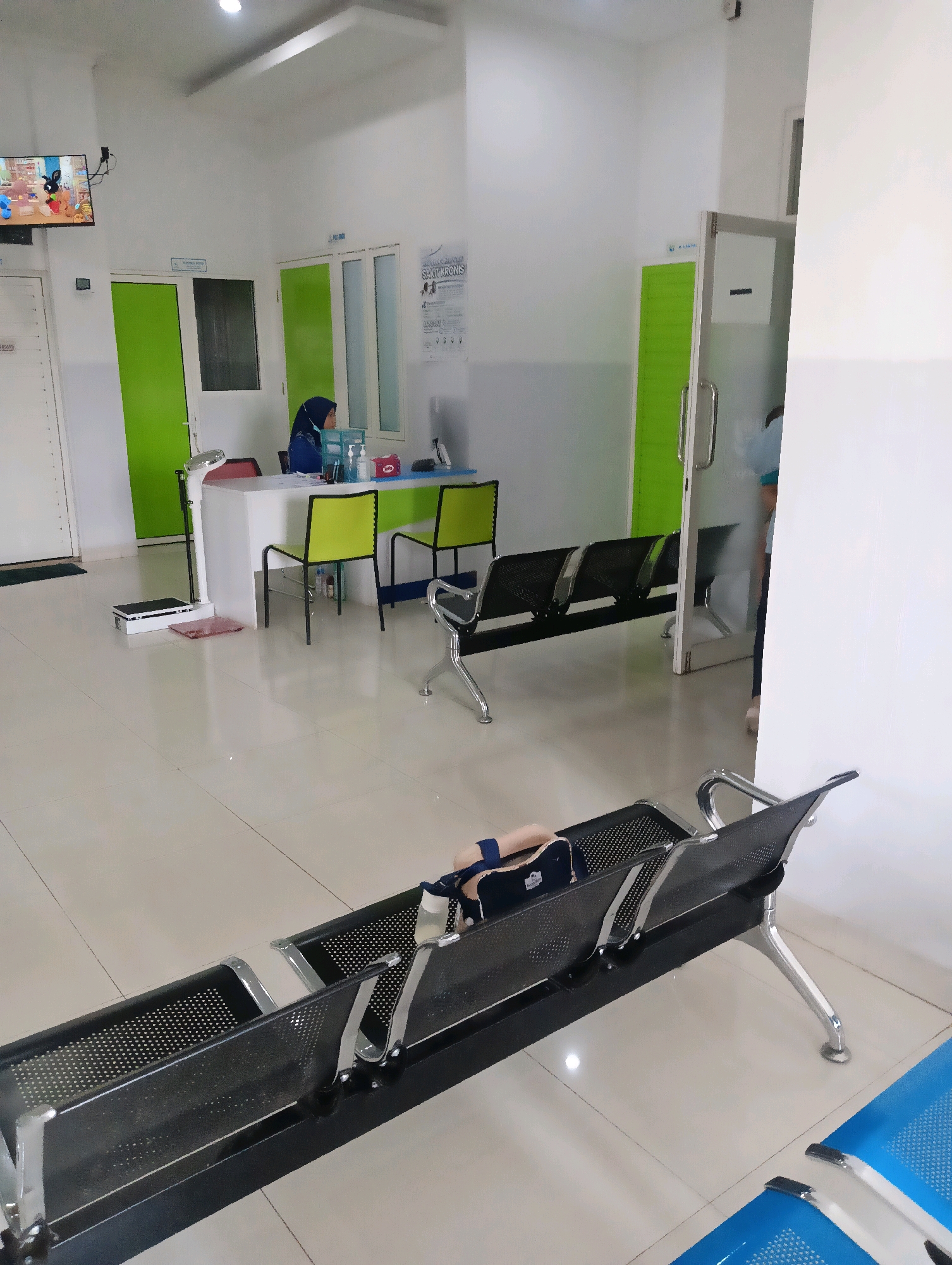 The image shows an interior scene, likely a medical clinic, with a reception area and a waiting area. In the reception area, a woman in a hijab and medical attire is seated behind a white counter. On the counter, there are various items including a tissue box, hand sanitizer, and what appears to be medical supplies. Adjacent to the reception desk is a digital scale. To the left of the reception desk, there are two doors, one of which is partially visible and painted green. A television screen is mounted on the wall to the left, displaying an animated scene.
The waiting area consists of rows of black perforated metal benches with chrome legs. In the foreground, a pair of legs wearing blue socks and black shoes are visible resting on one of the benches, suggesting a person is seated there, though they are mostly out of frame. To the right of the reception area, a glass-paneled door leads into another room, and a person in blue scrubs is partially visible inside. The floor is tiled with large, light-colored square tiles. The walls are painted white, with some sections in a light grey color. There are also two bright green doors visible in the background, one of which appears to be a clinic room. The lighting in the room is bright, suggesting overhead fluorescent lights. The overall environment appears clean and orderly. There are no specific visual cues to confirm the location as Purwareja, Indonesia, other than the prompt's assertion.