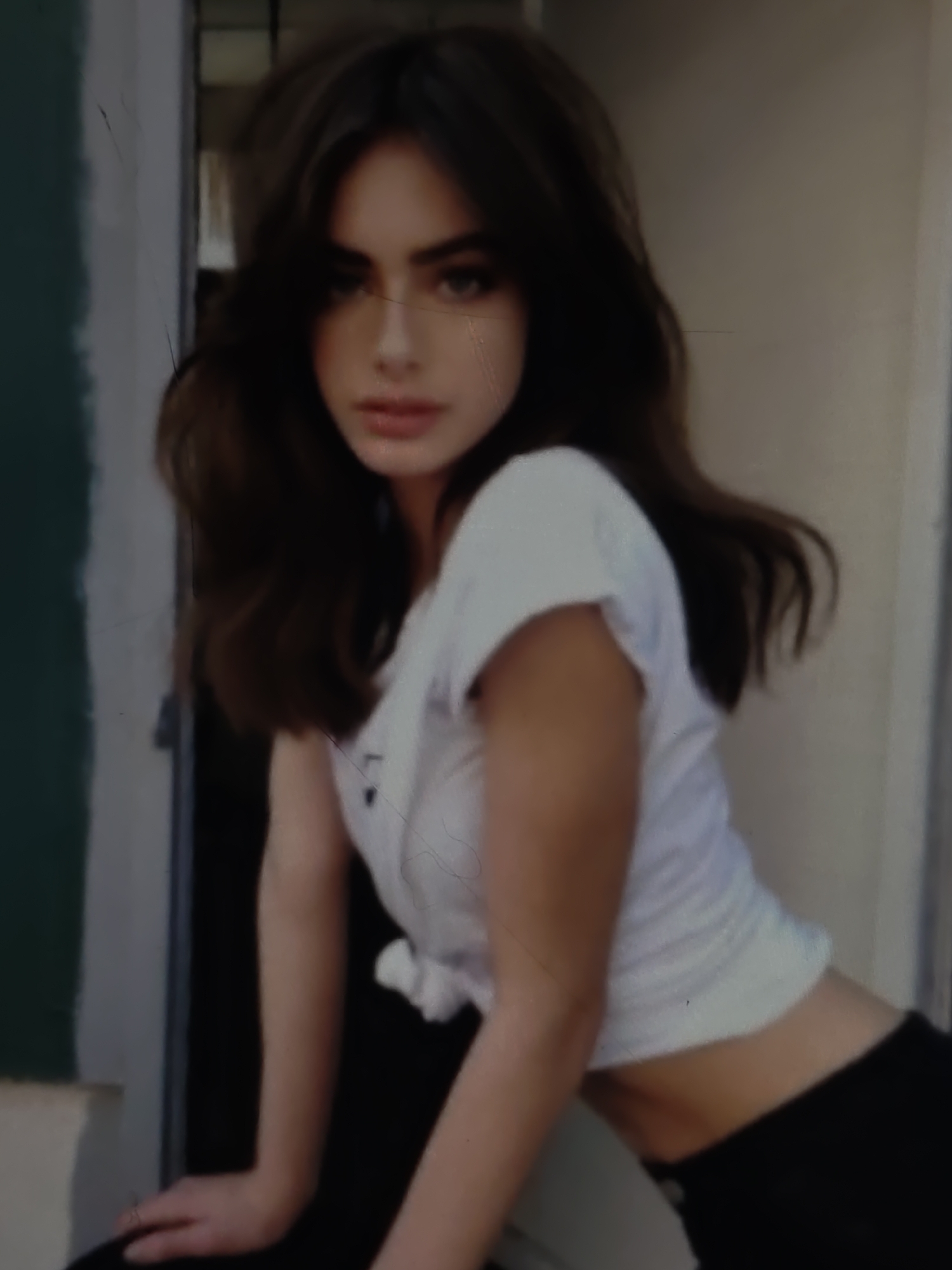 A young woman with long, dark, wavy hair is pictured from the chest up, leaning forward. She is wearing a white t-shirt tied at the waist, exposing her midriff, and black pants. Her gaze is directed towards the viewer with a neutral expression. The background consists of a blurred interior or exterior space with vertical lines suggesting a doorway or partition. There are subtle lines and a slightly grainy texture to the image, indicating it may be a photograph. The scene does not provide any specific indicators of its location within the city of Kurganinsk, Russia.