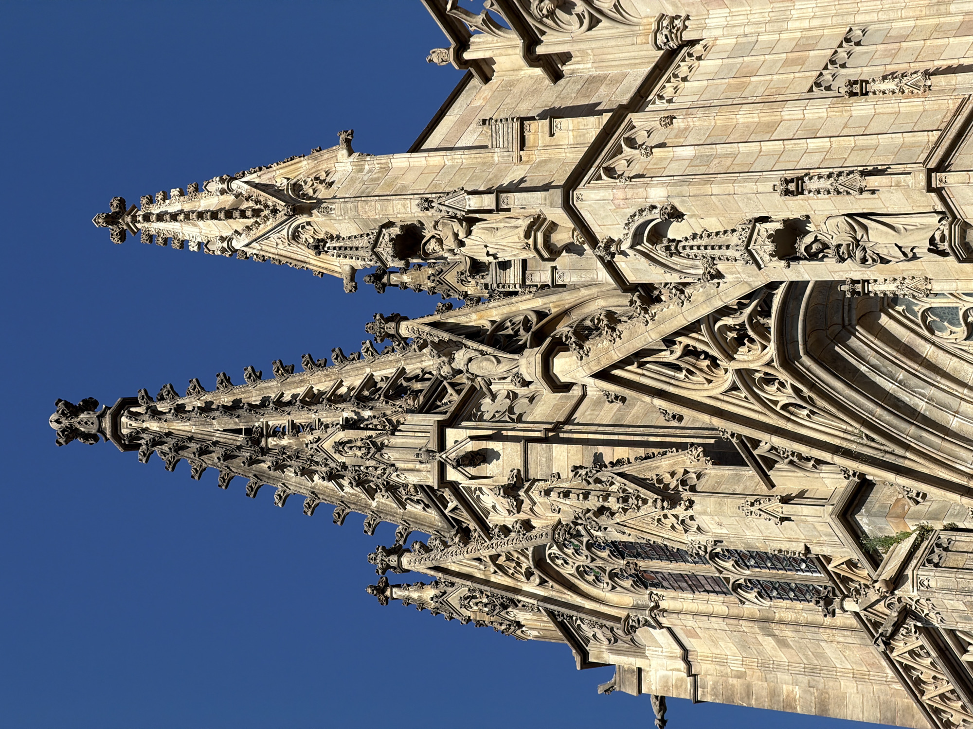 This is a low-angle, close-up, outdoor photograph of the upper portions of the Barcelona Cathedral in Barcelona, Spain. The architectural style is Gothic, characterized by soaring spires adorned with intricate stonework, gargoyles, and statuary. Two prominent spires dominate the frame, reaching towards a clear, deep blue sky. The stone of the cathedral is a warm, light brown, and shows signs of age and weathering. The details of the facade are sharp and clear, highlighting the delicate carvings and architectural elements, including pointed arches, tracery, and statues of religious figures. Sunlight illuminates the right side of the structure, casting shadows that emphasize the three-dimensional nature of the carvings. There are no people visible in the image. The environment is solely the cathedral and the sky.