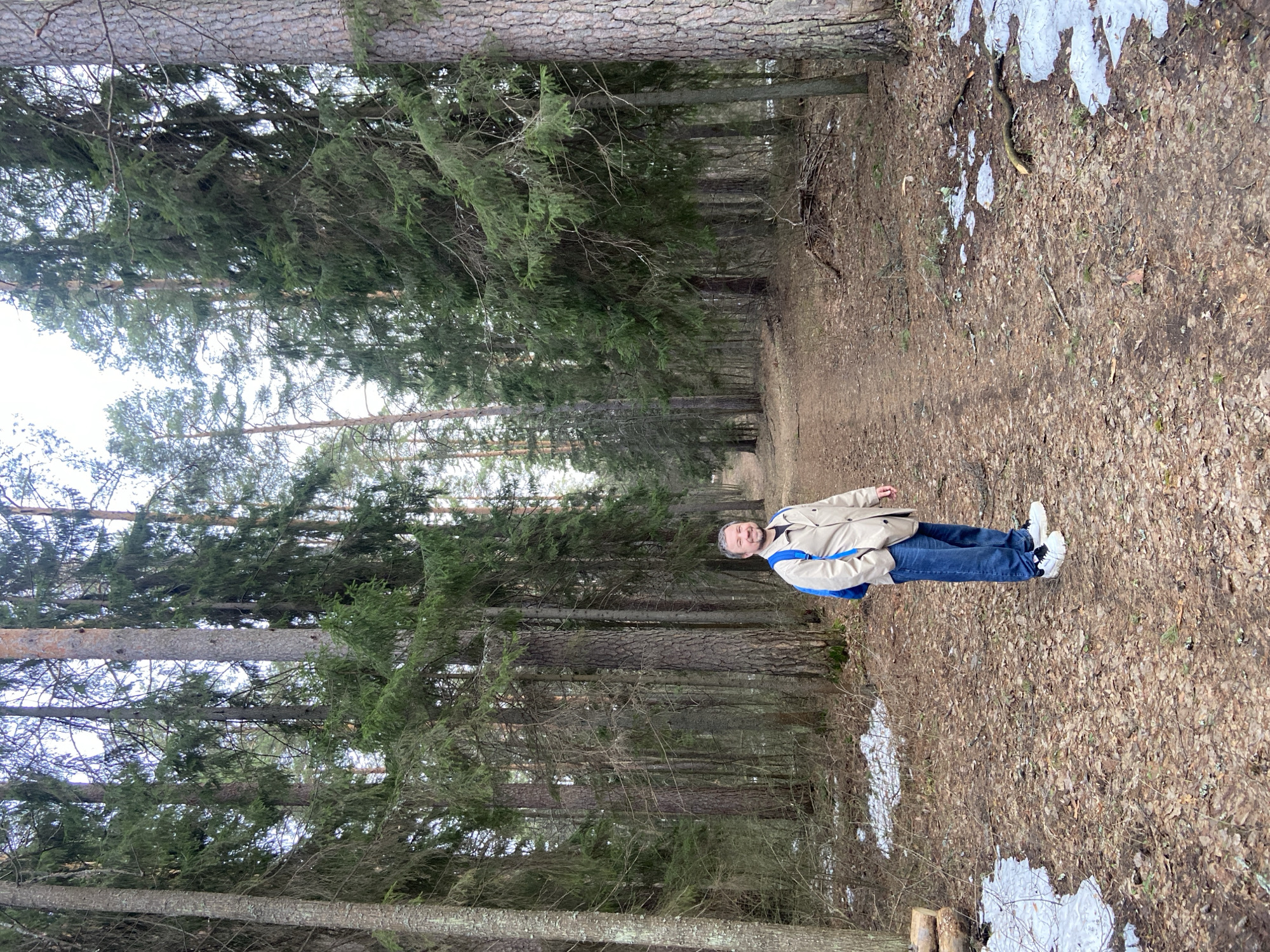A man stands on a woodland path. The path is covered in dead leaves and patches of snow and ice. Tall, straight pine trees line both sides of the path. The man is wearing a tan jacket with a blue backpack, blue jeans, and white sneakers. He is facing the camera with a slight smile. The sky is visible between the trees. The location is Noviy, Russia.