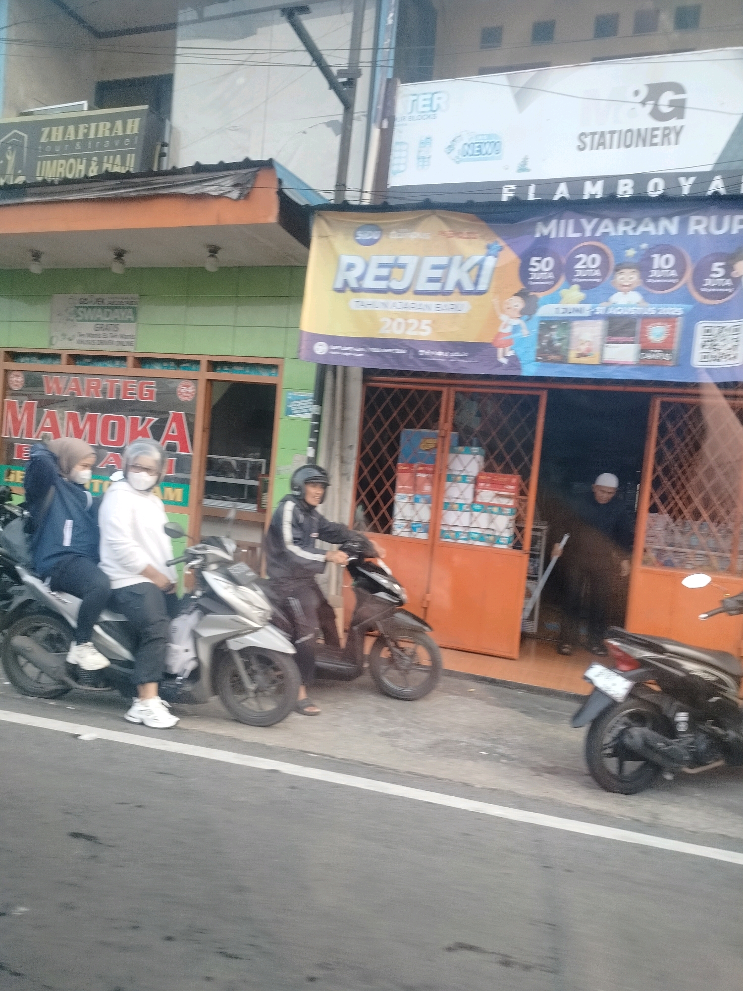 The image captures a street scene in Megamendung, Indonesia, focusing on the storefronts and the activity in front of them. On the left, a storefront displays a sign for "ZHAFIRAH TOUR & TRAVEL UMHROH & HAJJ" above an orange-trimmed awning. Below this, a green facade is visible with a sign that reads "SWADAYA." Further to the right, a large red sign on an orange storefront announces "WARTEG MAMOKA" with some text beneath it that is partially obscured. In front of the Mamoka Warteg, three individuals are on two motorcycles. Two women wearing hijabs and face masks are seated on a silver scooter, one in a dark blue jacket and the other in a white hoodie. A man wearing a black jacket with white stripes and a helmet is on a separate motorcycle to the right, appearing to be riding away.
Above the storefronts, signs indicate commercial businesses. One sign, partially visible at the top, appears to be for "STER" and "NEW!" with a logo that might be "PG STATIONERY." Another banner stretches across the scene above the orange storefronts, featuring text such as "REJEKI" and "MILYARAN RUP." This banner is yellow and purple, decorated with illustrations of children and circles containing numbers and the word "JUTA," suggesting a promotional event or lottery.
The environment appears to be a typical street in a commercial area, with paved road and sidewalks. Buildings are situated along the street, showcasing various businesses. The overall impression is one of a busy local commercial district.
