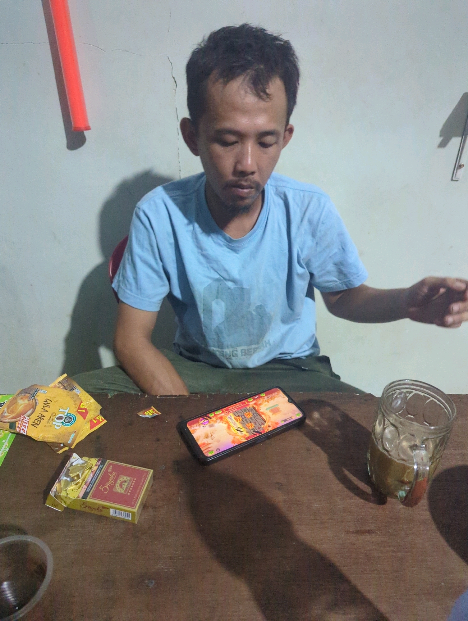 A man with short, dark hair and a light blue t-shirt sits at a wooden table. He is looking down at a smartphone in front of him. To his left are two yellow packets and a red and yellow cigarette pack. To his right is a glass filled with a light brown liquid and ice. A bright orange object, possibly a pool noodle, is visible on the wall behind him. The scene appears to be indoors, with a light-colored wall and a wooden table. The man's right hand is extended towards the right side of the frame. The image quality is average, with some graininess. There are no visible signs of distress or immediate danger. The environment suggests a casual setting.