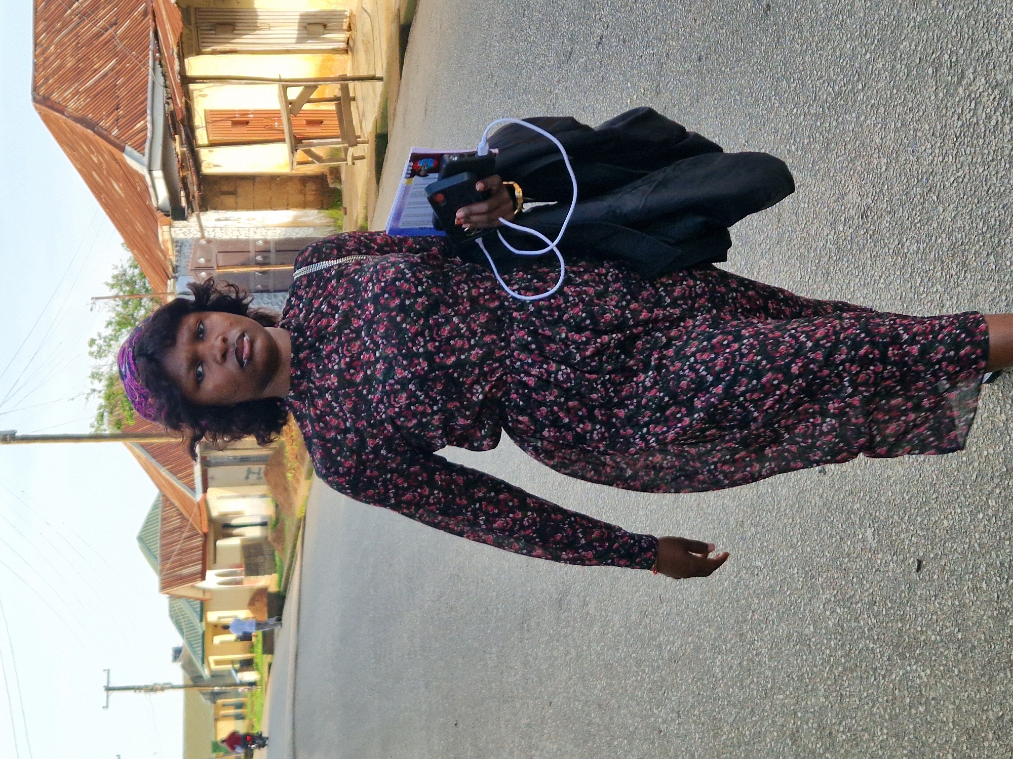 A woman with dark skin, wearing a floral dress and a purple headscarf, walks along an asphalt road in Kabong, Nigeria. She carries a black jacket over her left arm, and in her right hand, she holds a phone and what appears to be a book or folder, with a white charging cable attached. The woman is looking directly at the camera. In the background, there are single-story buildings with corrugated iron roofs and a few trees. A utility pole and power lines are visible in the distance, and a motorcycle is approaching from the left. The sun is shining, casting faint shadows.