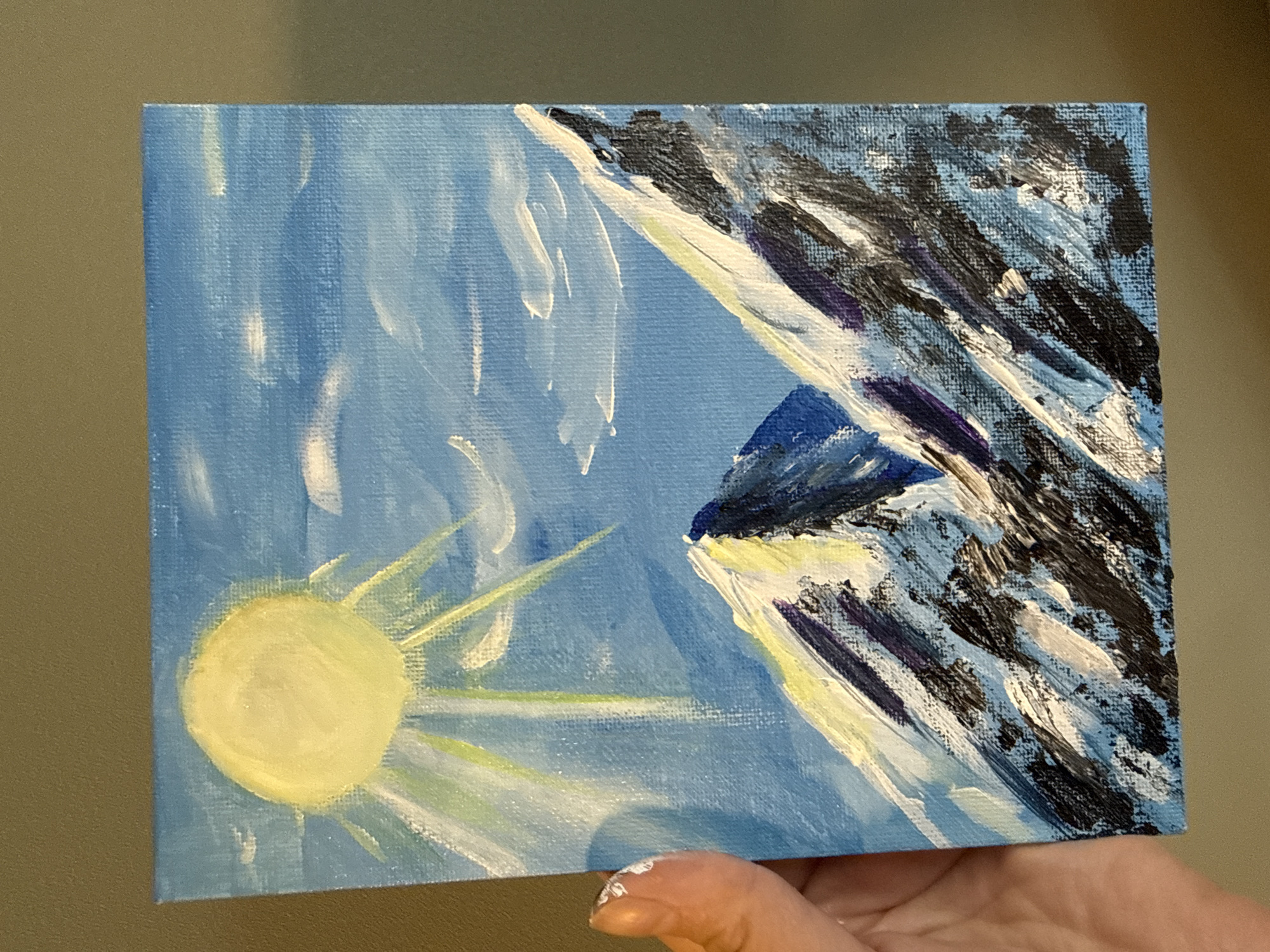 The image displays a small, rectangular canvas painting of a mountainous landscape under a bright sun. The sky is a vibrant blue with scattered white clouds. A large, yellow sun with rays of light, some tinged with green, dominates the upper left portion of the canvas. Two snow-capped mountains are depicted in the foreground, rendered with thick, impasto brushstrokes. The mountains are textured with shades of white, black, and deep blue, with highlights of yellow and purple. The painting is held by a person's left hand, whose fingers are visible on the left edge of the canvas, with a small amount of white paint on the thumbnail. The background behind the painting is a solid, muted green-brown color, likely a wall. There is no discernible information in the image to suggest its location in Cheboksary, Russia.