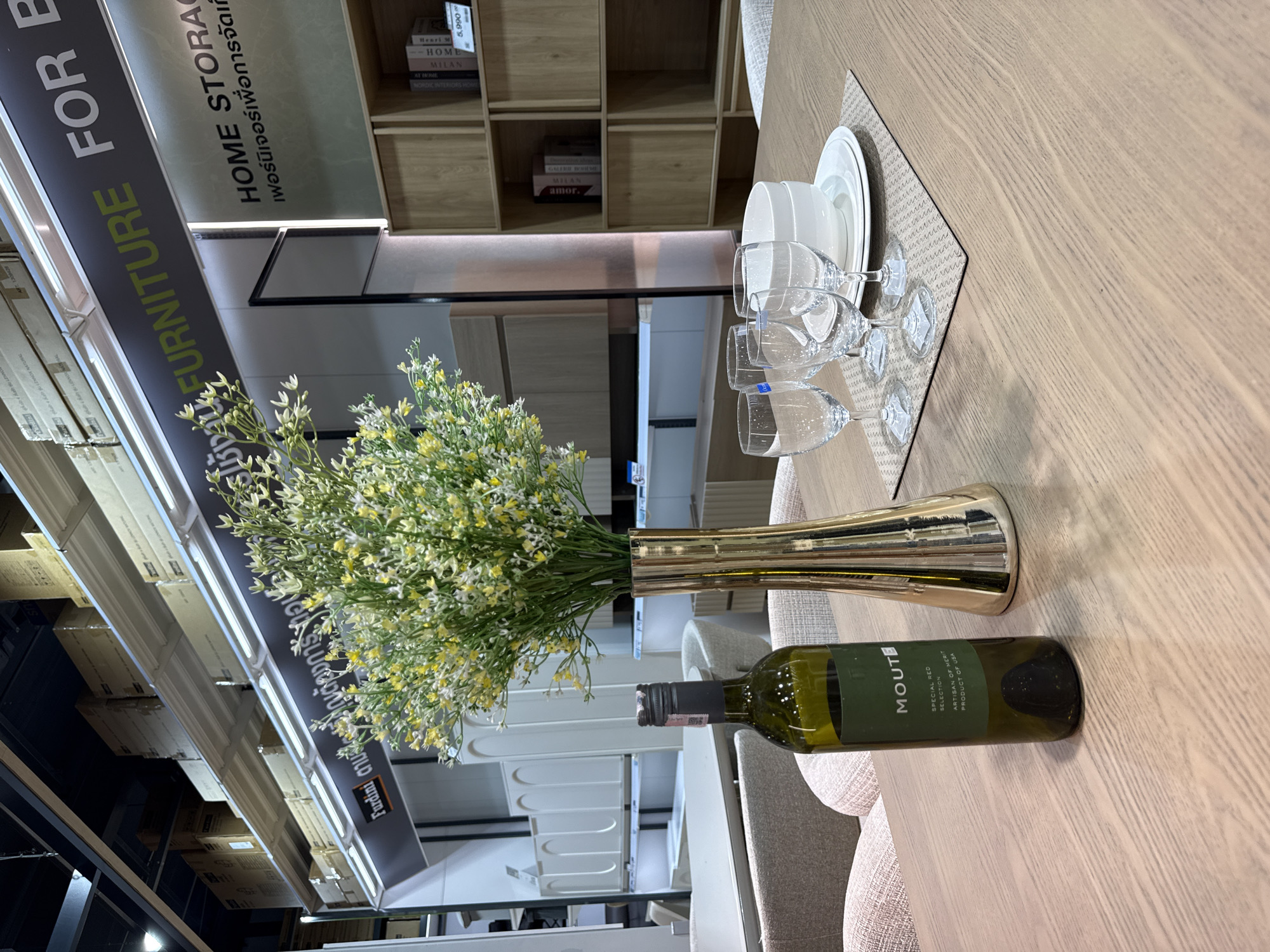 The image depicts an indoor scene within a furniture store in Baan Chaweng Noi, Thailand. The focus is on a wooden dining table set for a meal. On the table, there is a dark green bottle of wine labeled "Moutte Special Red Selection Product of USA," a gold vase filled with a large bouquet of small white and yellow flowers, and four clear stemmed wine glasses. In front of the glasses, a white placemat supports a stack of two white bowls and a white plate. In the background, shelves displaying boxed merchandise and furniture are visible. The environment appears to be a retail space designed to showcase home furnishings. There are no people visible in the image.