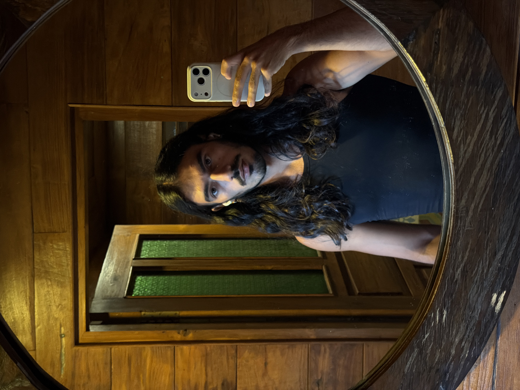 A person with long dark hair and a beard is taking a selfie in a round mirror. The person is wearing a black tank top. The phone is white with multiple camera lenses. The mirror is hanging on a wooden wall in what appears to be a bathroom or small room. In the mirror's reflection, a wooden door with green glass panes is visible. The lighting in the room is warm and slightly dim. The location is inferred to be in North Kuta, Indonesia, based on the environment and architecture.