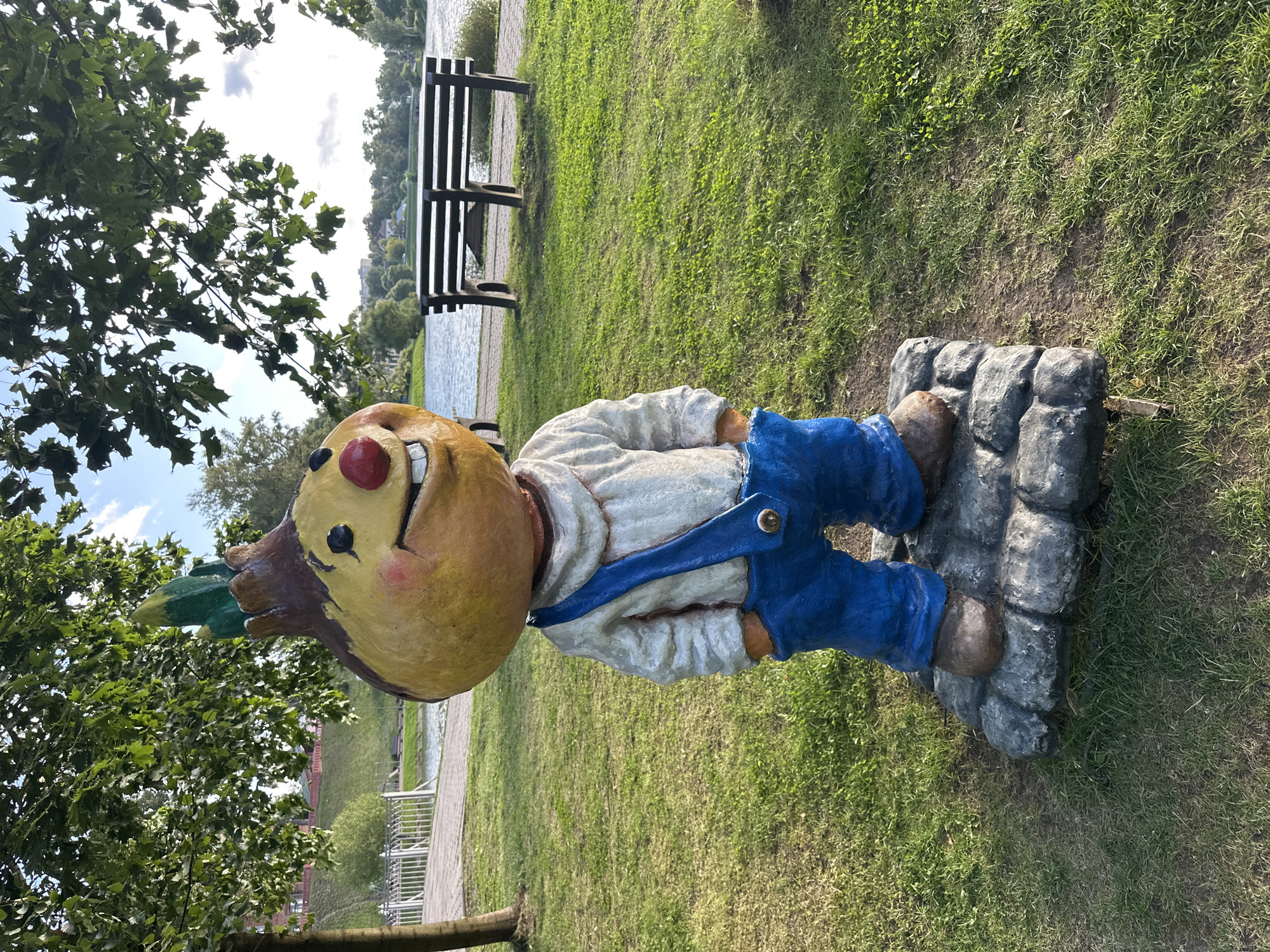 A statue of a character resembling an onion in blue overalls and a white shirt stands on a cobblestone base in a park. The character has a large yellow head with a red nose and a wide smile, and a green sprout on top. The statue is positioned in front of a body of water, likely a river or lake, with a wooden bench visible on the opposite bank. Trees with green leaves shade the area, and the ground is covered in grass. The background shows a cityscape with buildings and a bridge. This scene is located in the city of Sergiyev Posad, Russia.