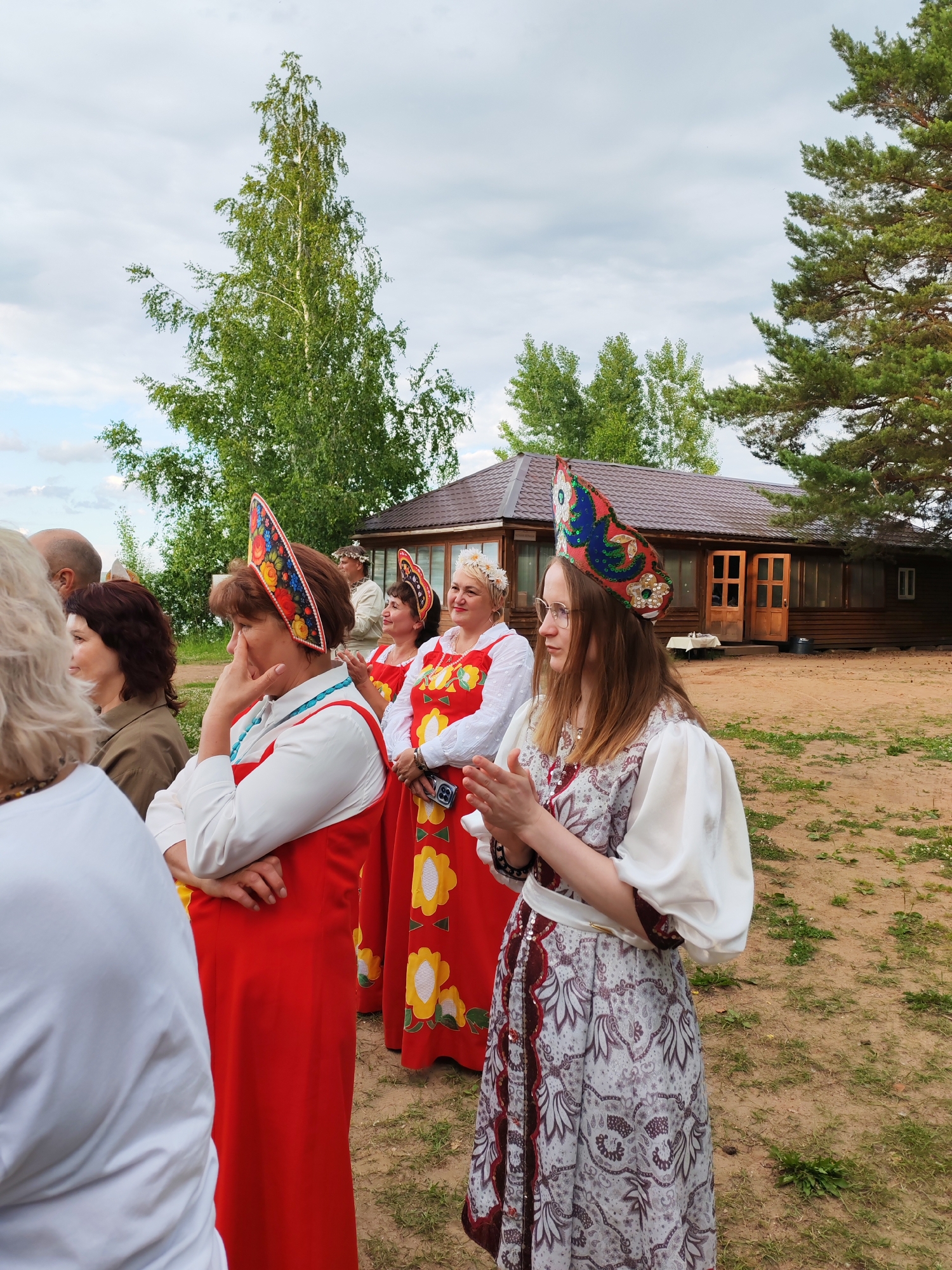 An outdoor scene at the ethnographic park of the Chaikovsky State Museum-Reserve in Chaikovsky, Russia, features a group of people, primarily women, dressed in traditional Russian folk costumes. In the foreground, a woman on the right wears a long, patterned dress with voluminous white sleeves and a decorative headdress. To her left, another woman is clad in a bright red dress with a white blouse and a similar ornate headdress, her hand touching her face. Behind them, other women in similar attire stand in a line. A wooden building with a brown roof and windows is visible in the background, set against a backdrop of trees and an overcast sky. The ground is sandy and uneven, with patches of grass.