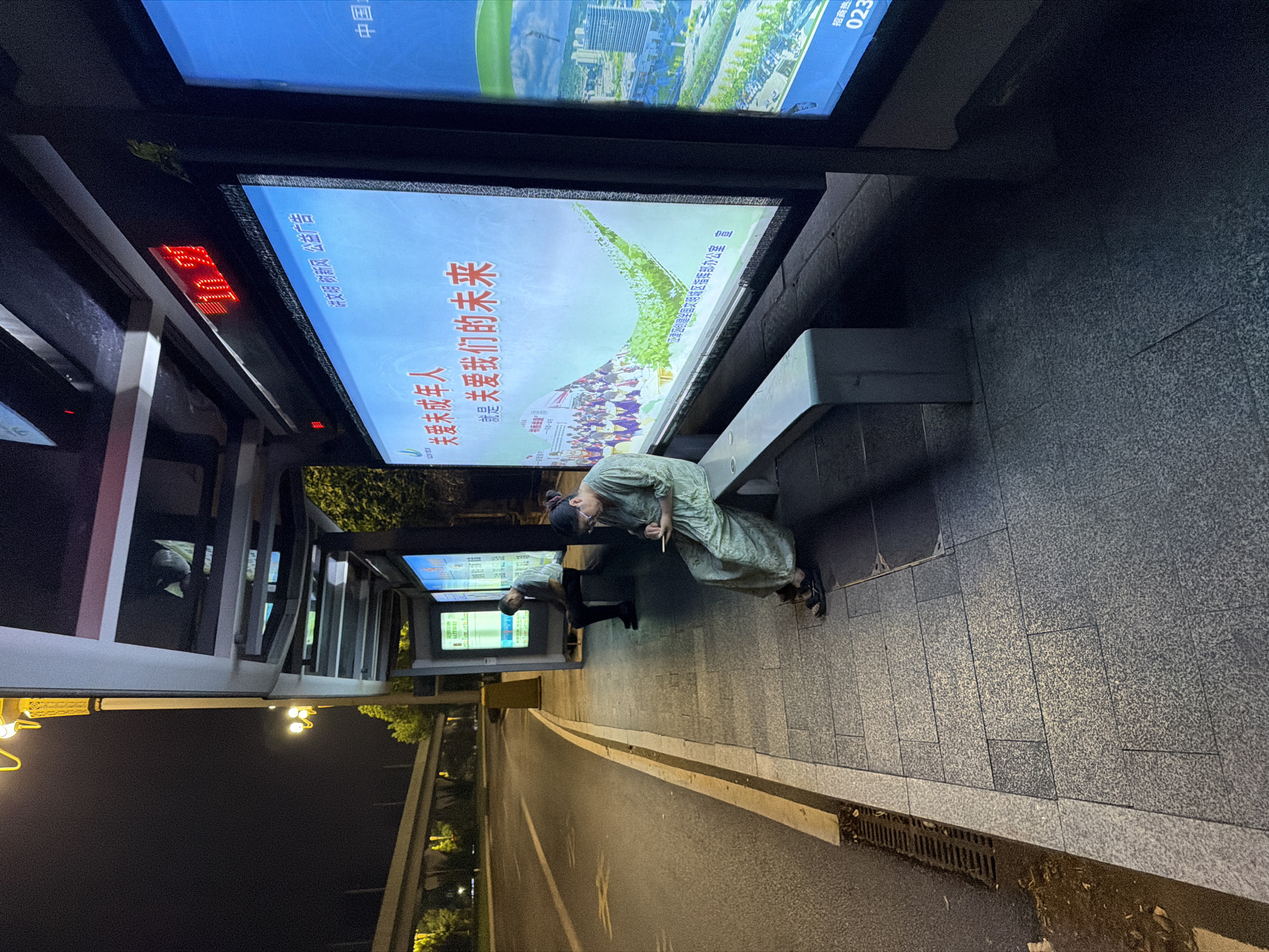 A night-time exterior shot captures a bus stop on an empty street in Jiangjin, China. A woman in a green patterned outfit sits on a bench, engrossed in her phone. Further down the bench, a man in a light-colored shirt is seated. Behind them, large digital billboards display bright advertisements. The bus stop is well-lit by overhead streetlights and the illuminated signs. The street and sidewalk are wet, suggesting recent rain. The scene is quiet and calm, with no vehicles or other pedestrians visible.