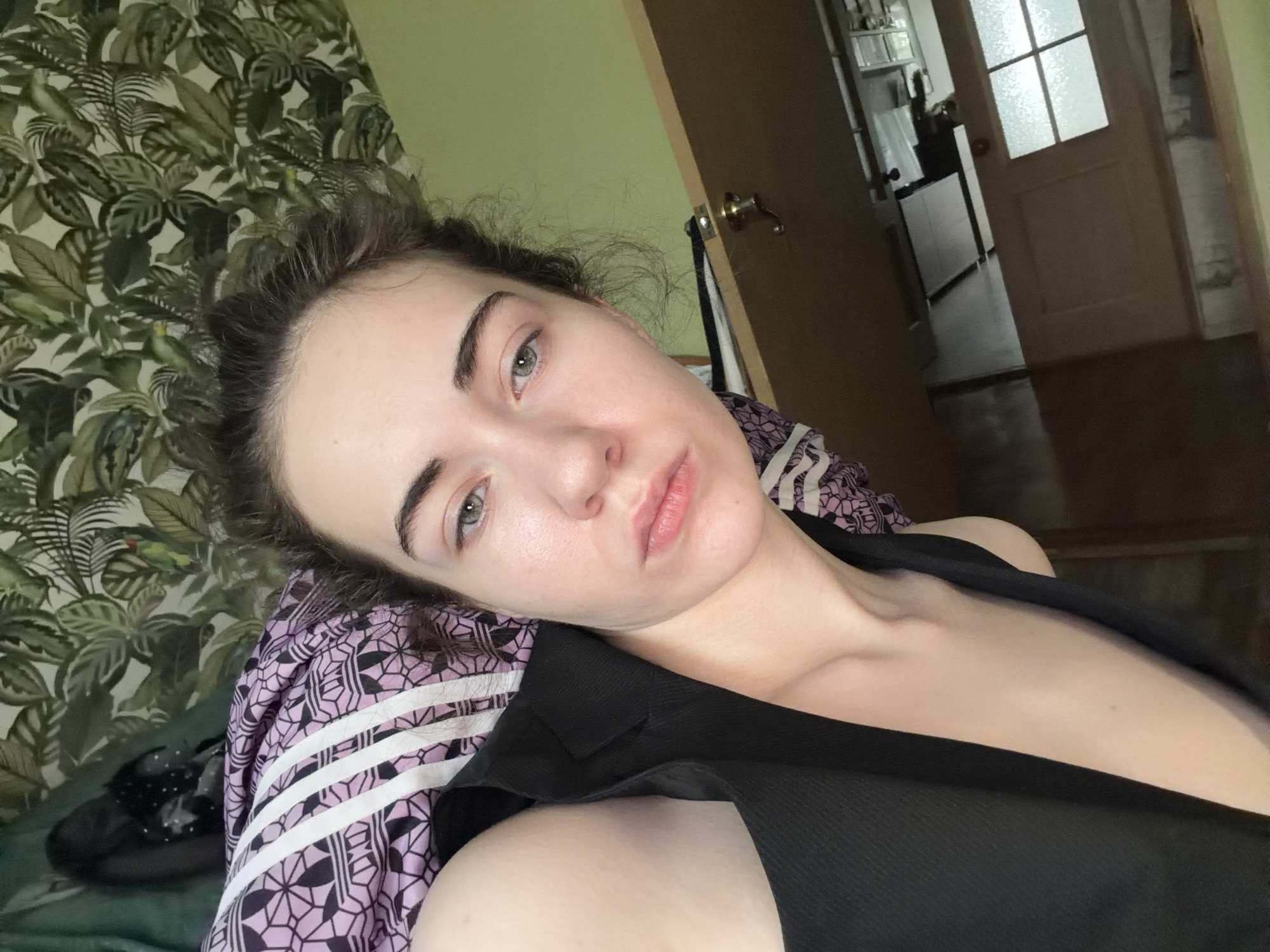 A young woman with light skin, fair complexion, and light green eyes is lying down, her head resting on a purple and white patterned pillow and blanket. She has dark brown hair that is slightly messy and pulled back. She is wearing a black top. The background includes a wall with green leaf-patterned wallpaper, a door, and what appears to be a kitchen or hallway. The lighting is dim. The setting is indoors, likely a residential room. This image was taken in Zavodoukovsk, Russia.