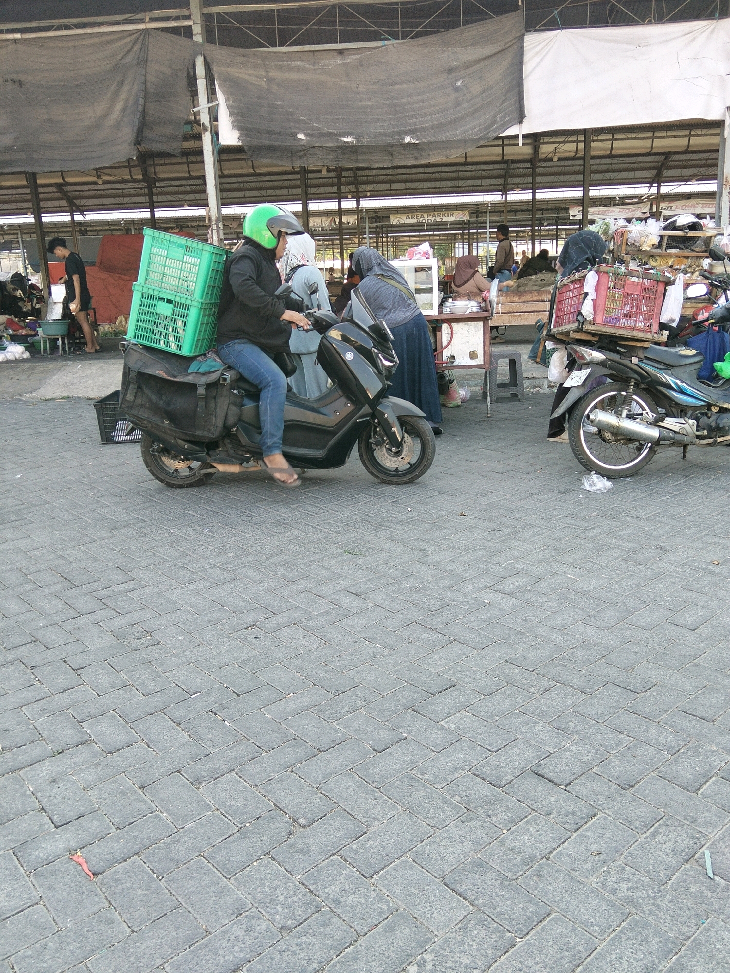 In Banyudono, Indonesia, a person wearing a green helmet and blue jeans rides a black scooter. The scooter has a large black bag attached to the back, with a green crate on top of the bag. The scooter is parked on a grey brick pathway. In the background, a market is visible with various stalls and people. Other motorcycles are also parked around the market.