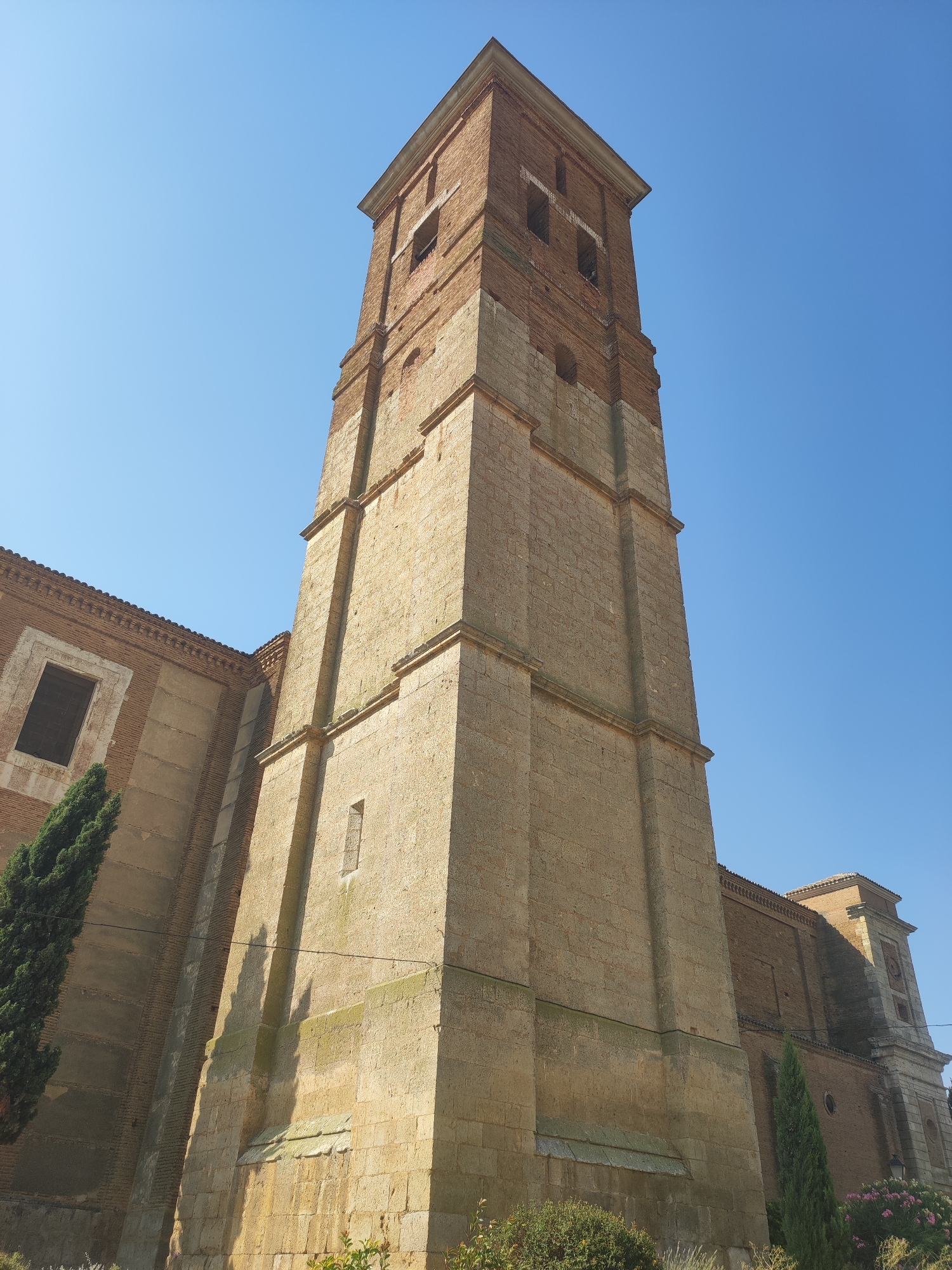 The image shows a low-angle view of a tall, weathered stone and brick bell tower of a church against a clear blue sky. The tower is square in its lower section, constructed from large, rough-hewn light brown stones, with horizontal banding. Above this, the tower transitions to red brick construction, featuring arched openings for bells at the top. The main body of the church, to the left, is also brick with some stone detailing and a large, dark-paned window. The church has a tiled roof. A tall, slender evergreen tree stands to the left of the tower, and other greenery and some pink flowers are visible at the base of the structure. The sun casts shadows of the tree and some architectural features onto the tower. The location is identified as the church of San Miguel Arcángel in Autillo de Campos, Spain.