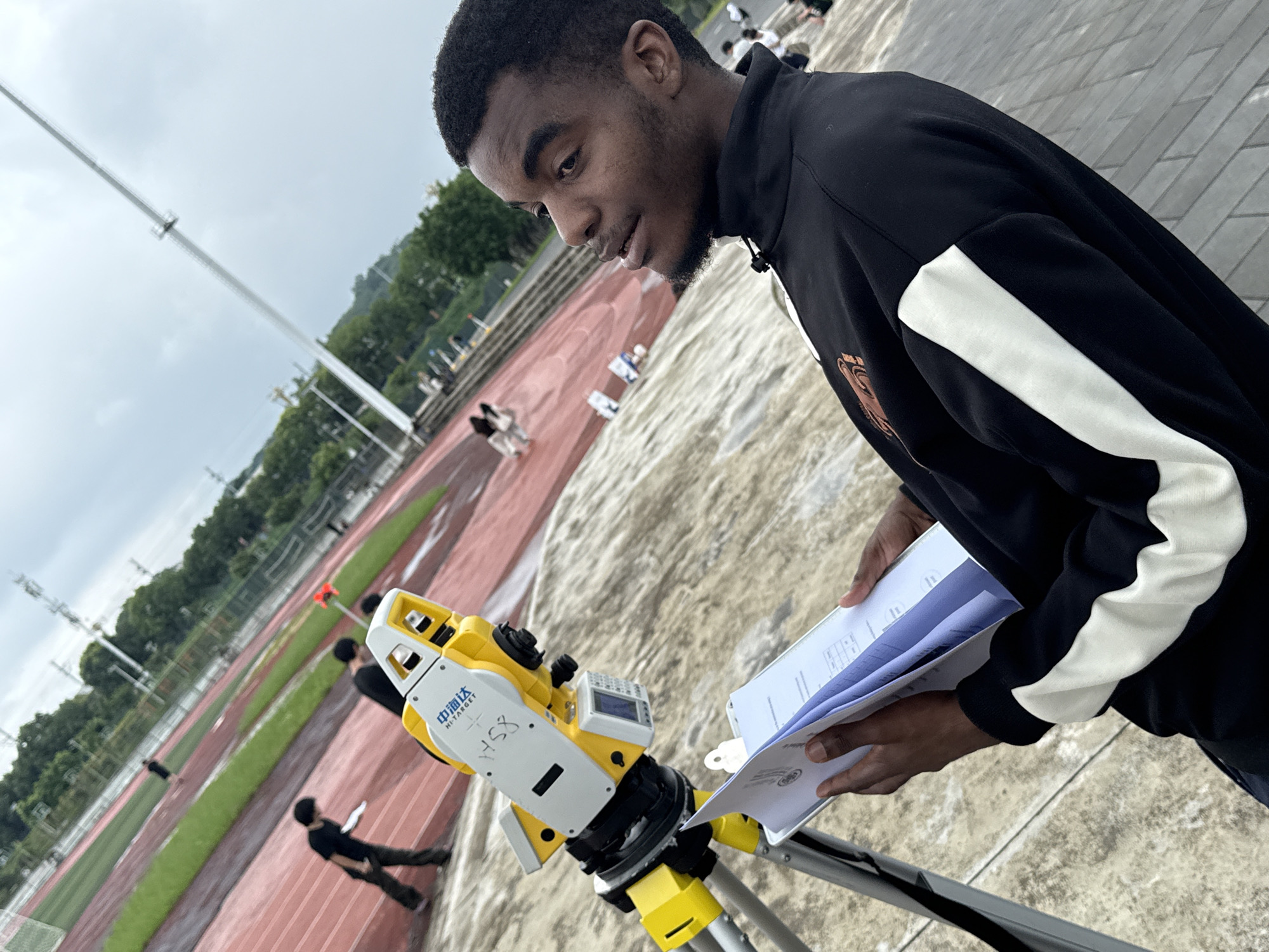 The image shows a young Black man holding a clipboard with papers while looking off to the right. He is wearing a black jacket with white stripes on the sleeves. In the foreground, a yellow and white surveying instrument, a total station, is mounted on a tripod. The instrument has Chinese characters and "HI-TARGET" printed on it. The background depicts a red running track with green infield, suggesting an outdoor sports facility, possibly a stadium or track and field. There are other people visible in the background, some appearing to be walking or running on the track. The sky is cloudy. The overall setting is consistent with an outdoor athletic complex in Jiangjin District, Chongqing, China, where such surveying activities might occur for infrastructure or event preparation.