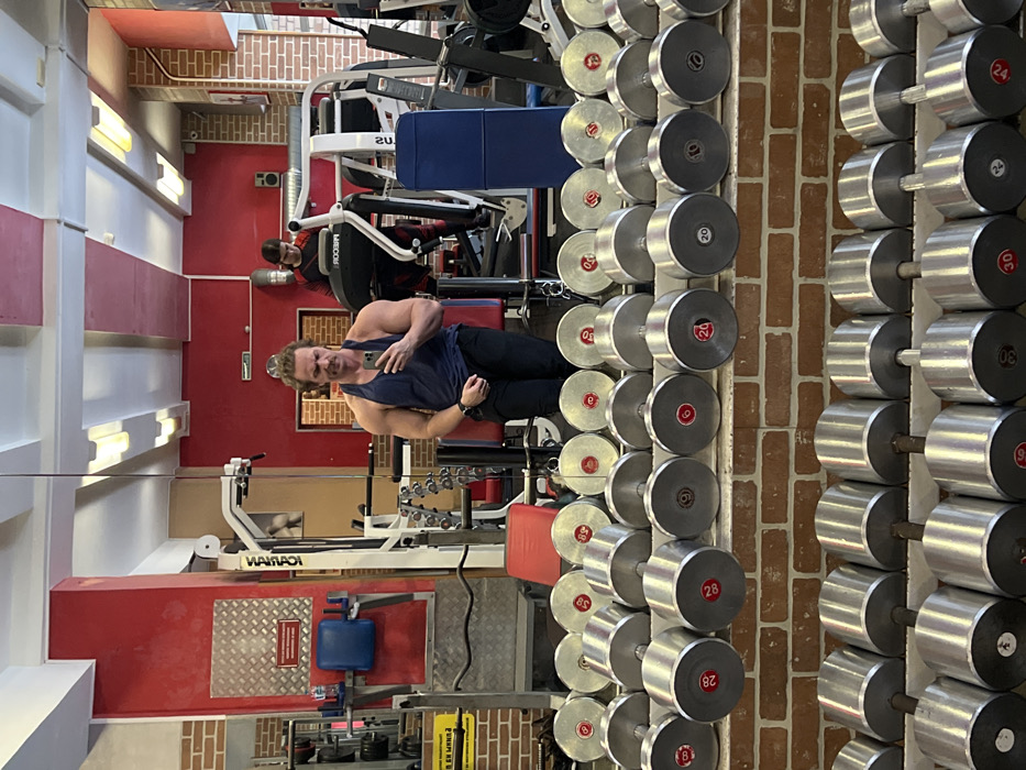 The image displays the interior of a gym. In the foreground, a multi-tiered rack, constructed from brick and metal, holds a symmetrical arrangement of numerous chrome-plated dumbbells. Visible weight markings on the dumbbells include 6, 8, 9, 10, 20, 24, 28, and 30 kilograms, often indicated with red numerals on their ends.
Reflected in a large mirror positioned behind the dumbbell rack, a muscular adult male is visible, appearing to be in his mid-thirties to forties. He wears a dark blue tank top and dark pants, has a mustache, and his hair is pulled back. He is holding a black smartphone in his right hand, angled towards himself, indicating he is taking a mirror selfie. A smartwatch is present on his left wrist.
Behind this male figure, also within the reflection, another individual is observed operating a grey Precor treadmill. This person is dressed in dark athletic wear with red patterns and is looking downwards.
The gym features red-painted walls, sections of exposed red brickwork, and white ceilings with rectangular fluorescent light fixtures. Red structural pillars are prominent, one featuring a diamond plate metal panel. Various other gym machines, including cable apparatuses with a "WARIAO" logo and a red-cushioned bench, are visible in the background. A white sign with a black arrow is present in the upper right background. There are no discernible markings, signs, or unique architectural features within the image that allow for the precise identification of its specific location or address within the city of Saint Petersburg, Russia.