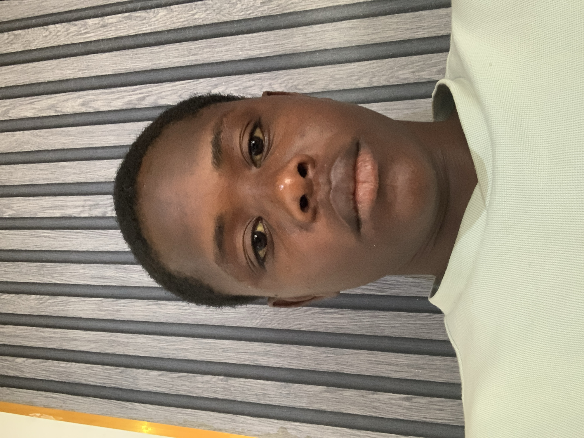 The image shows a close-up, head-and-shoulders portrait of a young person with dark skin and short dark hair. The subject is looking directly at the camera. They are wearing a pale green t-shirt with a textured fabric. The background consists of vertical, ribbed panels, alternating between a light grey wood grain pattern and a darker grey color. To the left of the background, a vertical orange element is partially visible. There are no discernible indications of the city of Jos, Nigeria, or any specific urban or environmental features within the image. The lighting appears to be from the front, illuminating the subject's face.