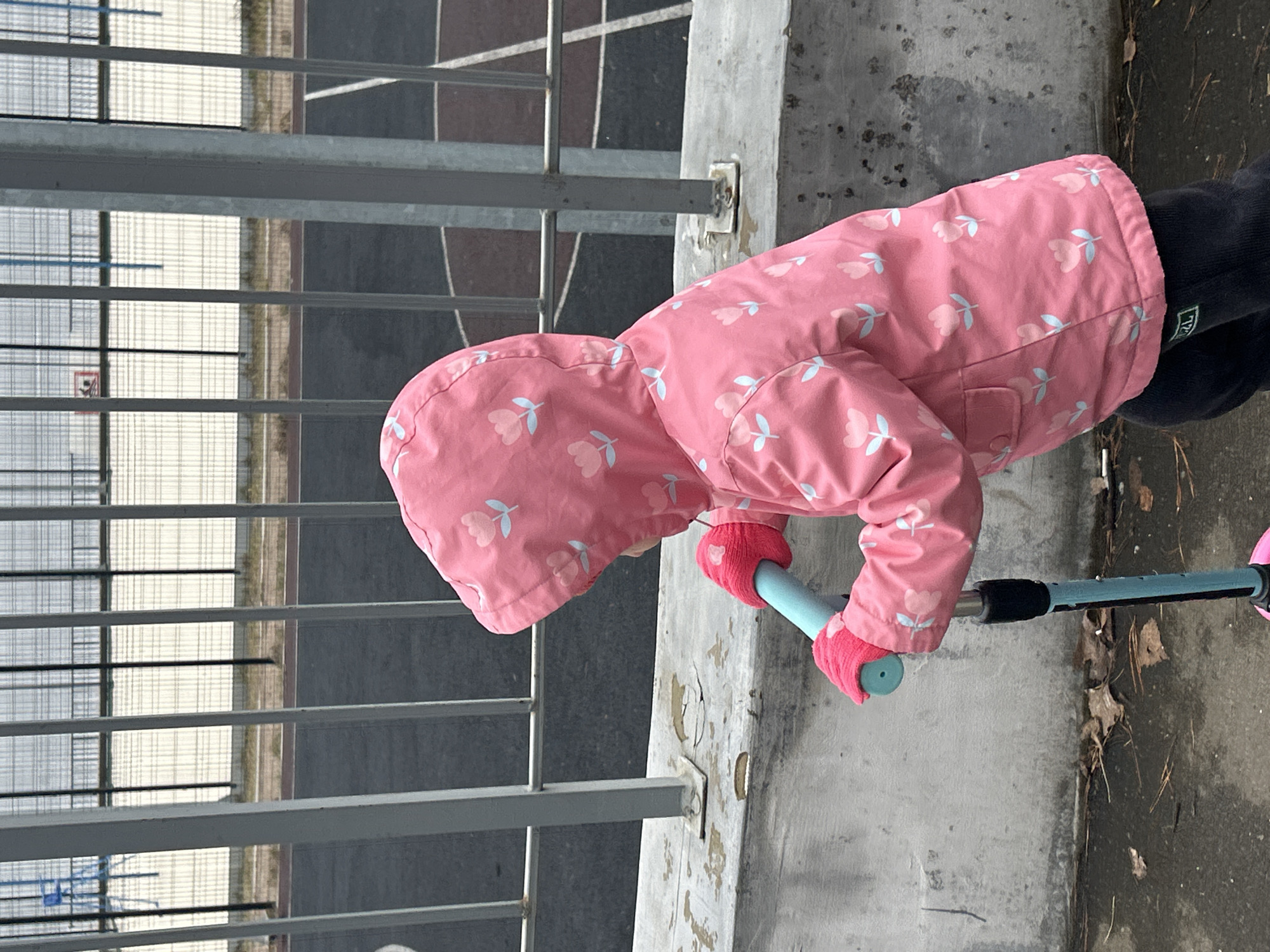 An outdoor scene in Chaikovsky, Russia, shows a young child from the back and side, engaged with a kick scooter. The child is wearing a vibrant pink hooded jacket with a repeating light blue and pink tulip pattern, dark pants, and bright pink gloves. Their left hand, covered by a pink glove, grasps the light blue handle of a scooter, which also features black and pink elements.
The child appears to be standing beside the scooter, possibly preparing to ride or having just paused. To the right of the child, a weathered concrete barrier or ledge shows signs of wear and minor damage. In the background, behind a multi-barred metal fence, a paved area stretches out, characterized by white line markings typical of a sports court or playground. A partial view of what seems to be a basketball hoop is visible in the distance. The presence of a jacket and gloves suggests cool weather conditions. A small green brand label, possibly "L.L.Bean," is visible on the outside of the child's left pant leg near the ankle. The overall setting appears to be a public outdoor recreational space during daytime.