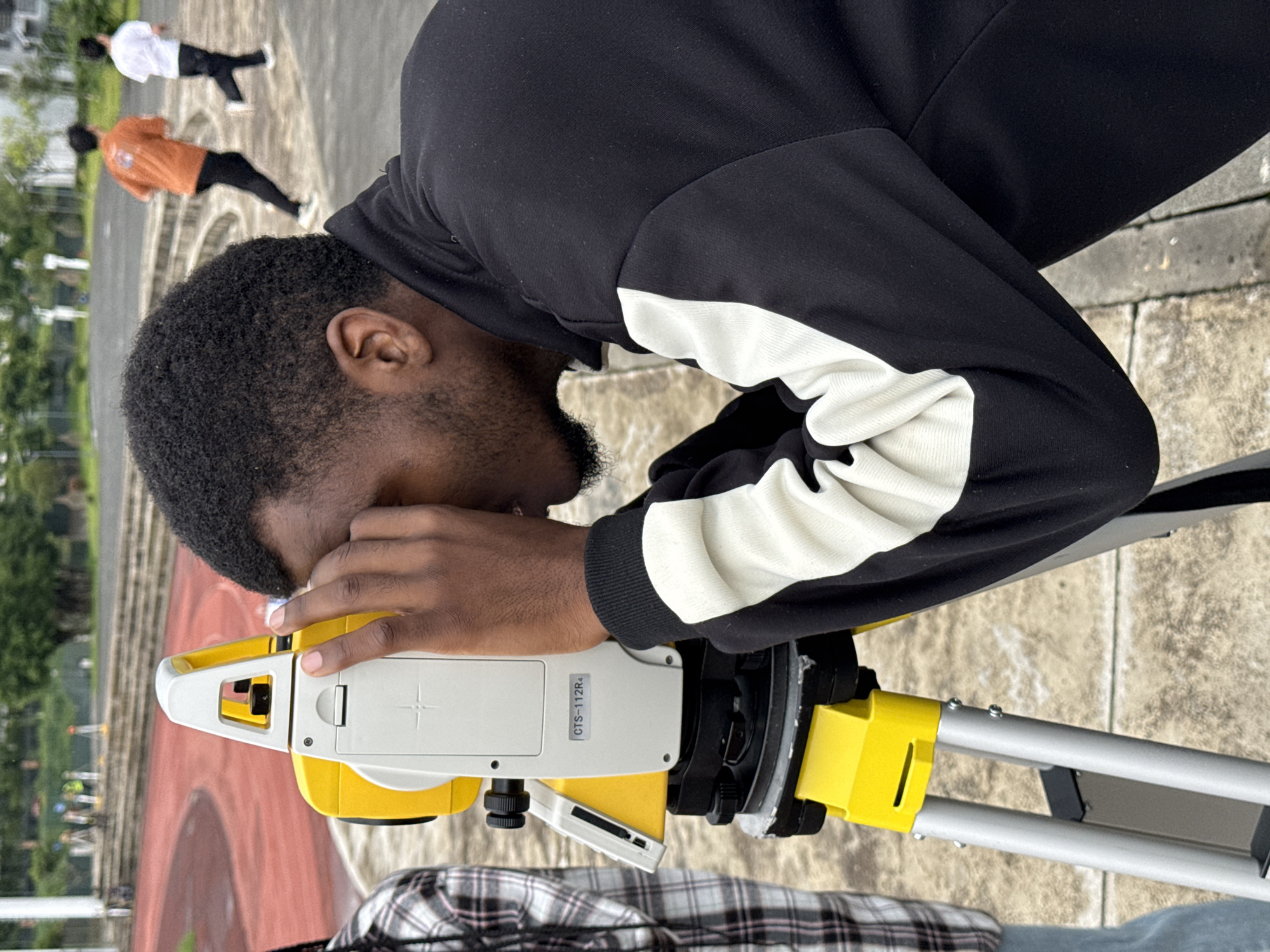 In the city of Jiangjin, China, a young Black man is shown in the foreground, looking through the eyepiece of a yellow and white surveying instrument, likely a theodolite or total station, which is mounted on a tripod. He is wearing a black jacket with white stripes on the sleeves. To his left, a person in a plaid shirt is partially visible. In the background, there are two individuals running on a track. The environment appears to be an outdoor recreational or educational area, possibly a campus or sports facility, with a red running track and tiered seating visible behind the surveying equipment. The ground surface is paved with what looks like concrete or stone pavers.