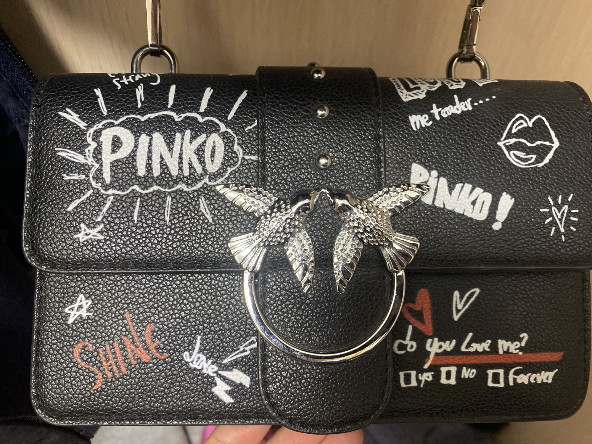 A black Pinko handbag is depicted in a close-up shot, likely indoors in Saint Petersburg, Russia. The handbag is made of black pebbled leather and features a distinctive graffiti-style design with white and red markings. The prominent clasp on the front is silver-toned and shaped like two stylized swallows pecking at each other over a ring.
Various handwritten-style texts and drawings adorn the bag's surface. On the top left, "PINKO" is written within a white speech bubble with radiating lines. Further to the right, "me tender...." is written in white, followed by a drawing of white lips. Another "PINKO!" in white is visible below the lips.
On the bottom left, the word "SHINE" is written in red, with a partial "Love" in white next to it, accompanied by white lightning bolt drawings. On the bottom right, a playful "do you Love me?" is written in white, followed by checkboxes for "yes", "NO", and "Forever", with the "NO" box clearly checked in white. Red and white heart drawings are also scattered across this area.
The background appears to be a light brown or beige surface, possibly a shelf or a wall, with a hint of blue fabric on the far left. A hand is partially visible at the bottom, holding the bag. The overall impression is a stylish and edgy accessory.