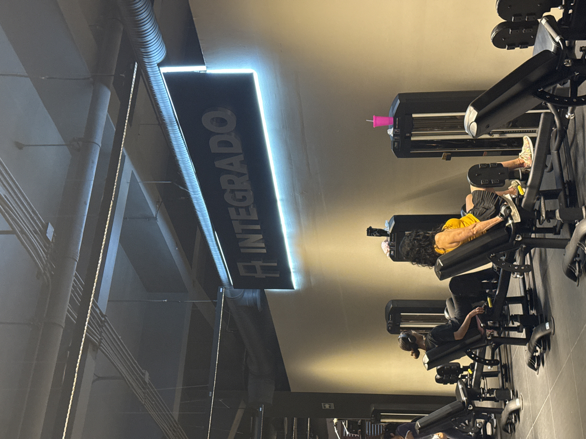 The image captures an indoor scene, likely within a fitness center in Puebla, Mexico, indicated by the hanging sign that reads "INTEGRADO." There are several people exercising on leg press machines in the foreground. These machines are uniformly black. The environment is well-lit, and the interior design includes exposed piping and ductwork on the ceiling. The scene appears to depict a typical exercise session with people engaging in leg workouts.