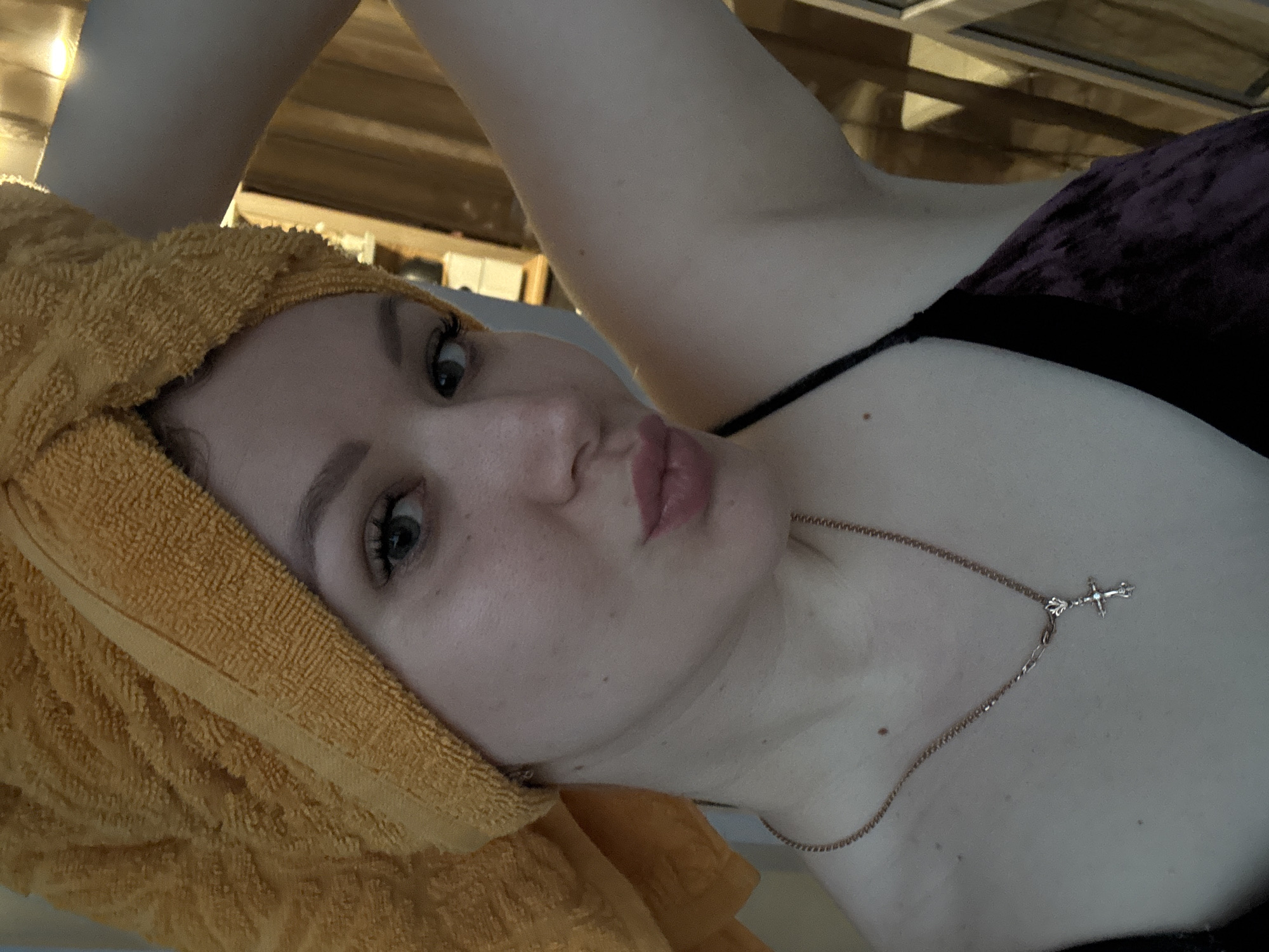 A woman with fair skin and green eyes is seen from a close, low-angle perspective. She is wearing a gold necklace with a cross pendant and a dark top. A yellow towel is wrapped around her head. The background shows a partially visible wooden structure, possibly a ceiling or wall. The photo appears to be taken indoors. There is no explicit information to confirm the location is in the city of Chaikovsky, Russia. No activities or interactions are depicted in the image other than the subject posing for the photograph.