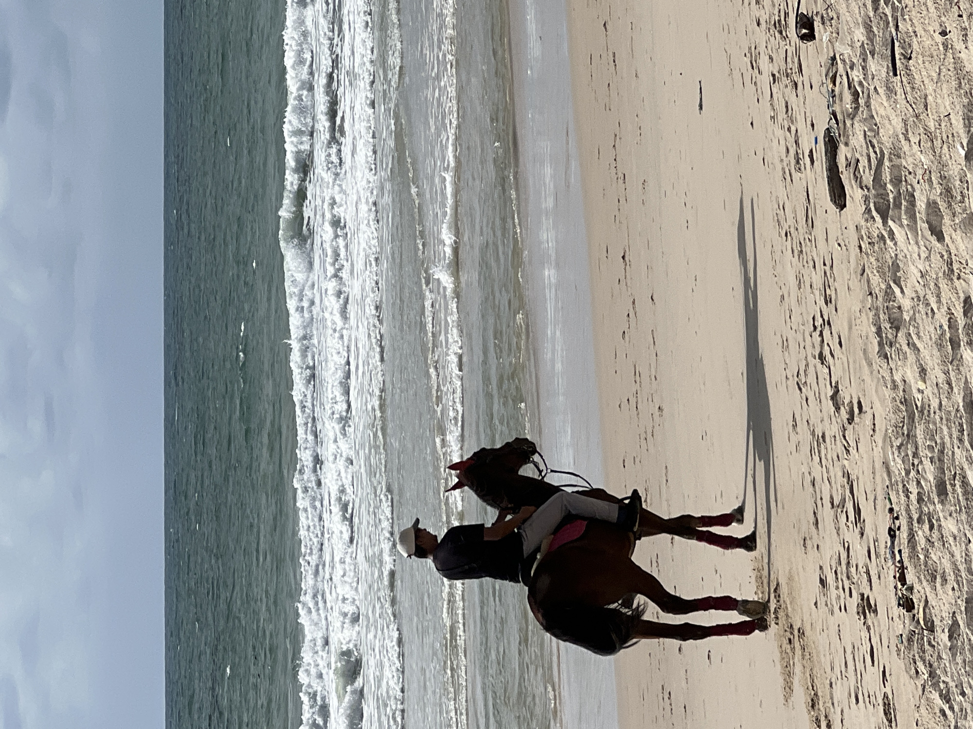 A person is riding a brown horse on a sandy beach. The horse is facing left, and the rider is looking forward. The rider is wearing a black t-shirt, grey pants, and a white cap. The horse is wearing a pink saddle pad and red leg wraps. The ocean is in the background with waves breaking on the shore. The sky is cloudy. The scene appears to be in Choeng Thale, Thailand, a coastal area known for its beaches.