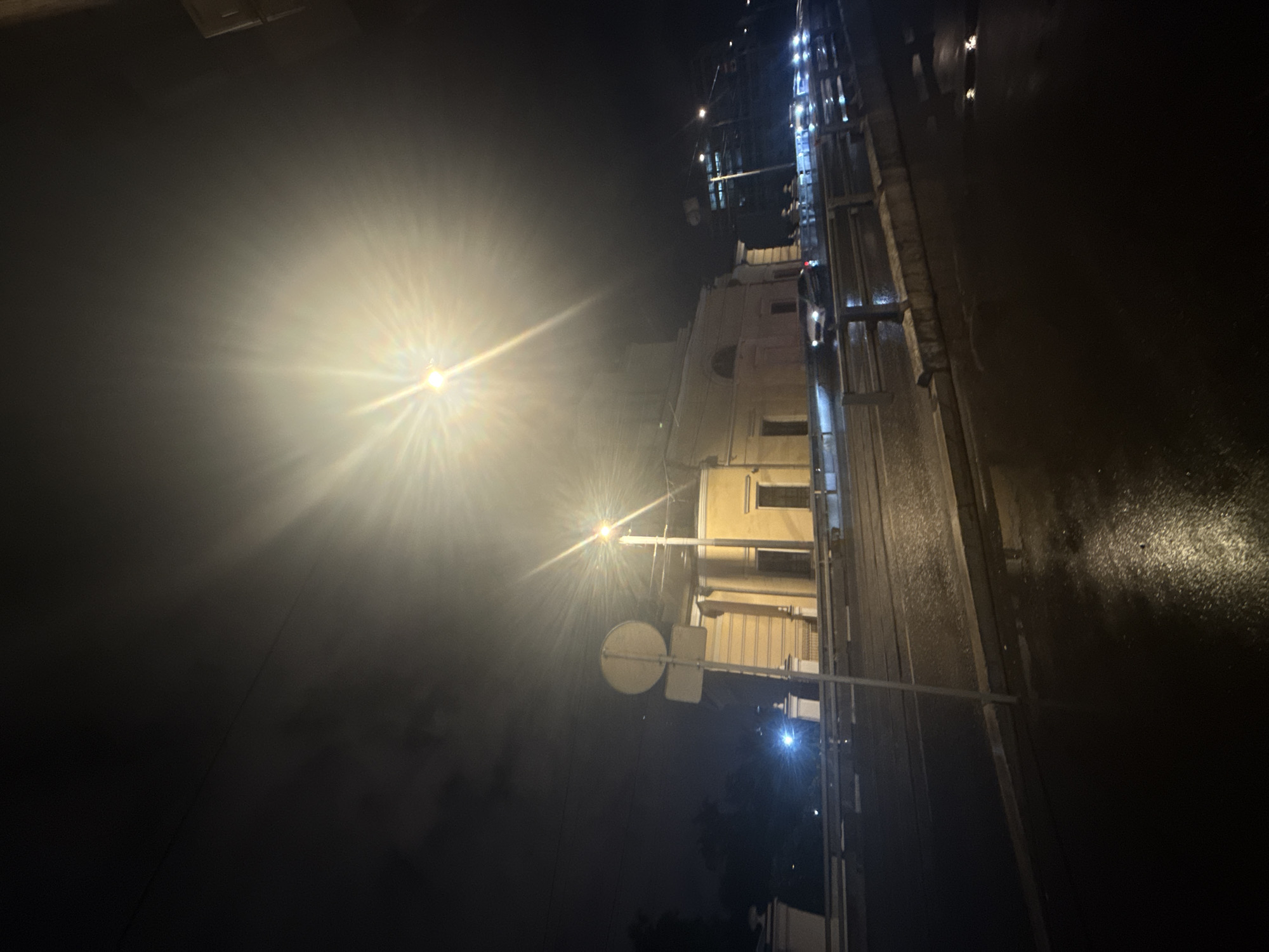 At night in Moscow, Russia, the image shows a street scene with a building, streetlights, and road. The lighting is harsh, with bright streetlights casting strong rays into the surrounding darkness. There appears to be a multi-story building illuminated with some windows visible. A road runs alongside the building, showing a wet surface with visible reflections. Traffic signals can be seen. The scene is dark and urban, with a focus on the contrast between artificial light and natural darkness.