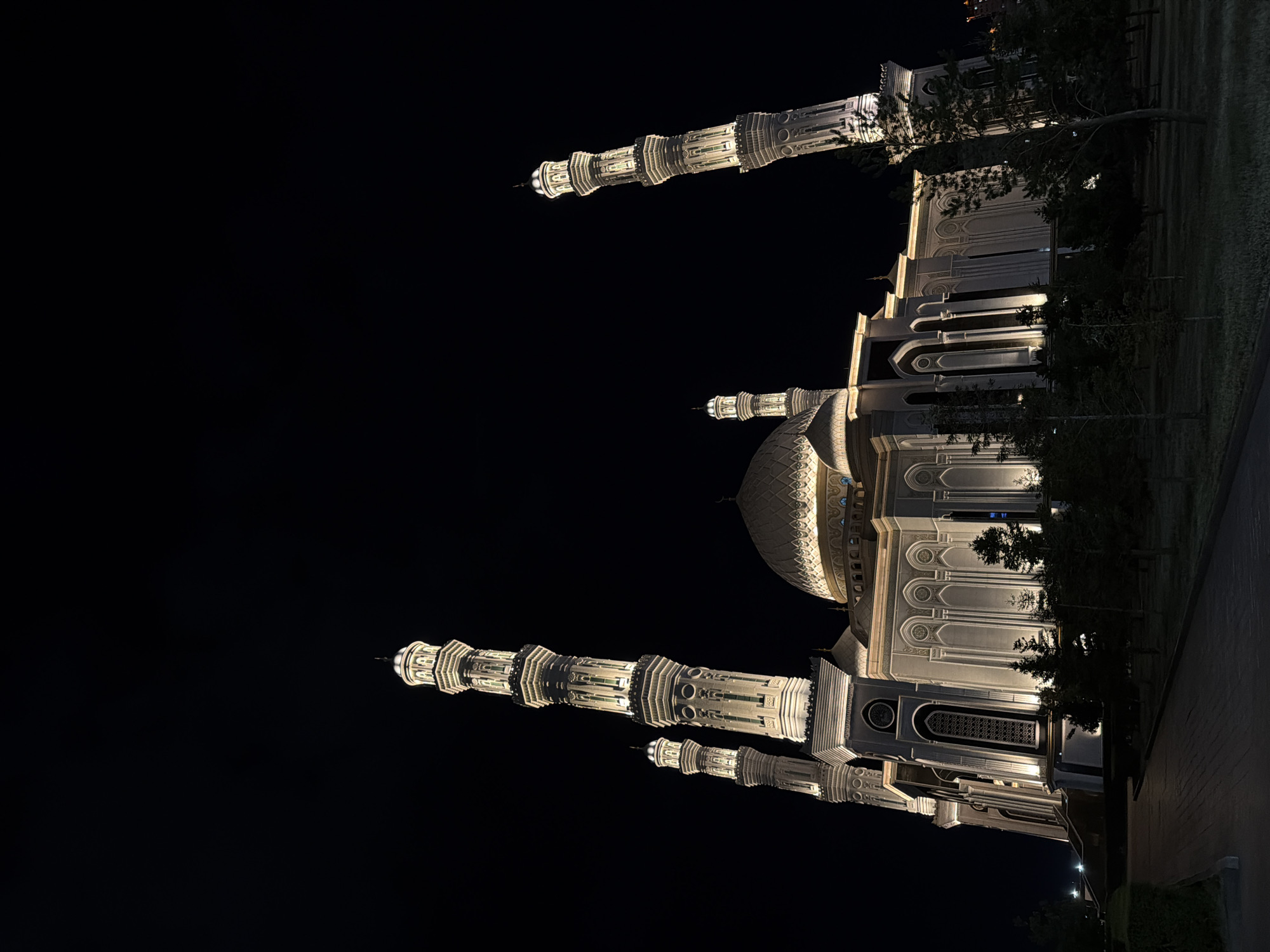 This media file depicts an illuminated mosque at night, likely in Astana, Kazakhstan, given the location context. The primary subject is the mosque's grand architecture, characterized by its intricate detailing and lighting. Prominent features include multiple tall, slender minarets topped with decorative finials and a large central dome. The building's facade showcases ornate carvings, arched windows, and symmetrical designs, all highlighted by warm artificial lighting that contrasts sharply with the dark night sky.
The scene is set outdoors, with the mosque grounds appearing to be a park or courtyard, featuring trees and pathways. The time of day is clearly night, as indicated by the dark sky and the artificial illumination of the structure. The weather appears clear, with no visible atmospheric disturbances. The overall mood conveyed is one of solemnity and architectural splendor, emphasizing the beauty of the mosque in the darkness. There are no people visible in the image. No visible text is present.