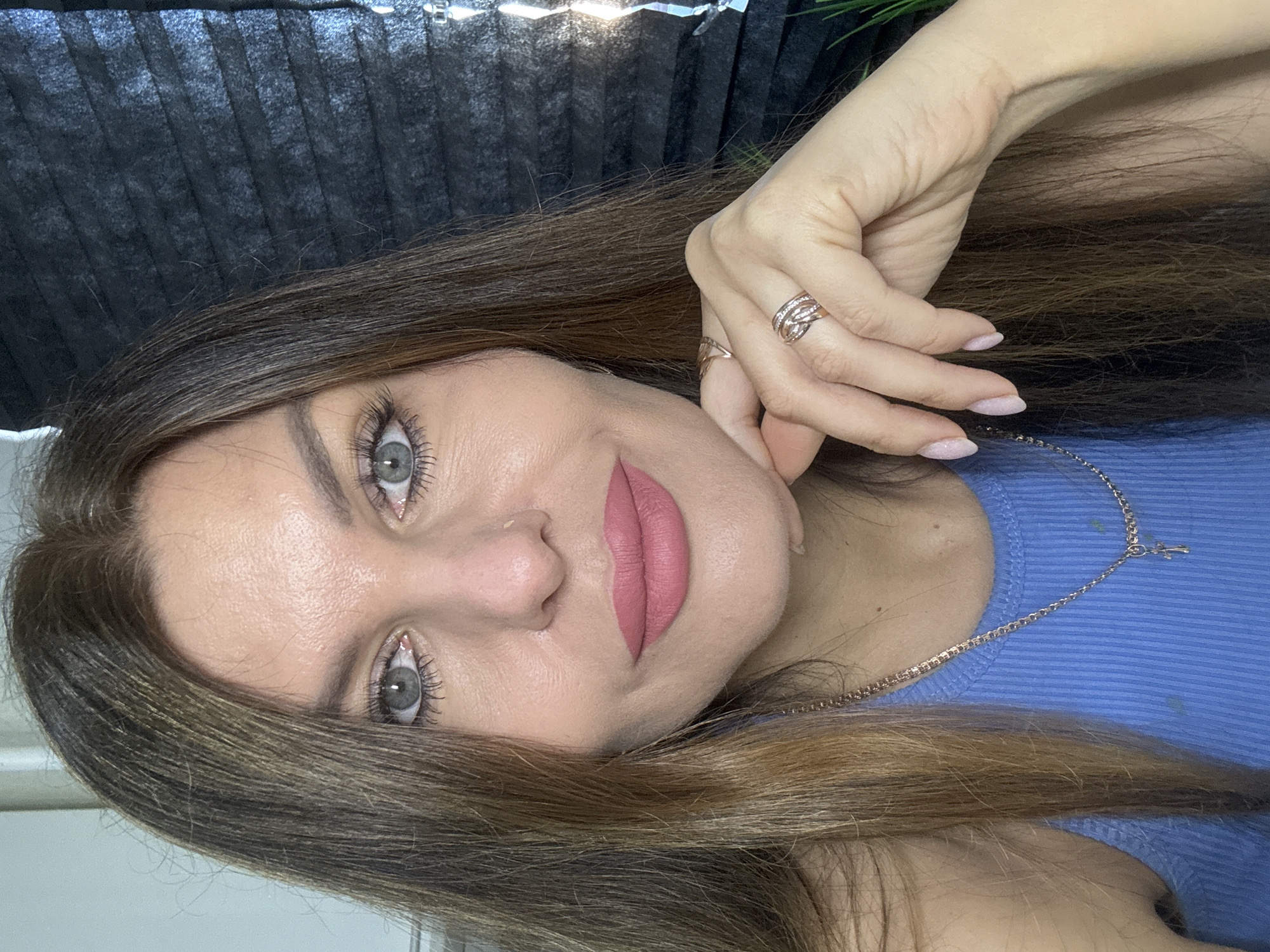 A close-up, angled portrait of a woman with light skin, grey eyes, and light brown hair with blonde highlights. She is wearing a blue ribbed top and a gold chain necklace with a small cross pendant. She has a nose ring and is wearing pink lipstick. She has two rings on her left hand, which is near her face. The background includes a dark, pleated material and some green foliage. There is no information available in the image to determine its location within Chaikovsky, Russia.
