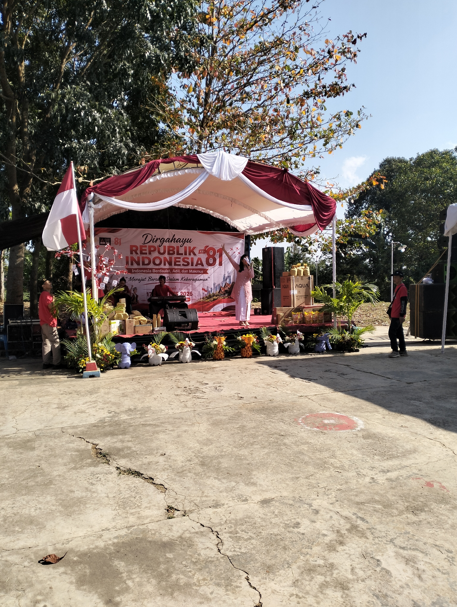 The image captures an outdoor stage setup for a celebration, likely in Semarang, Indonesia, given the Indonesian flag and banner. A red and white tent covers the stage, adorned with white fabric drapes. The banner behind the performers reads "Dirgahayu REPUBLIK INDONESIA KE 81" (Happy 81st Republic of Indonesia Day), with additional text below.
On the stage, a woman in a light pink outfit is singing into a microphone. To her right, a person is playing a keyboard, and another person is partially visible behind them. A variety of objects are arranged on the stage, including boxes of "AQUA" bottled water and bottles of cooking oil, as well as decorative plants and flower arrangements. Two individuals stand on the ground in front of the stage, one on the left in a red shirt and khaki pants, and another on the right in a red shirt and dark pants.
The stage is set on a paved outdoor area with visible cracks and some markings on the ground. In the background, lush green trees and a clear blue sky suggest a warm, sunny day. The overall scene depicts a festive event taking place outdoors.