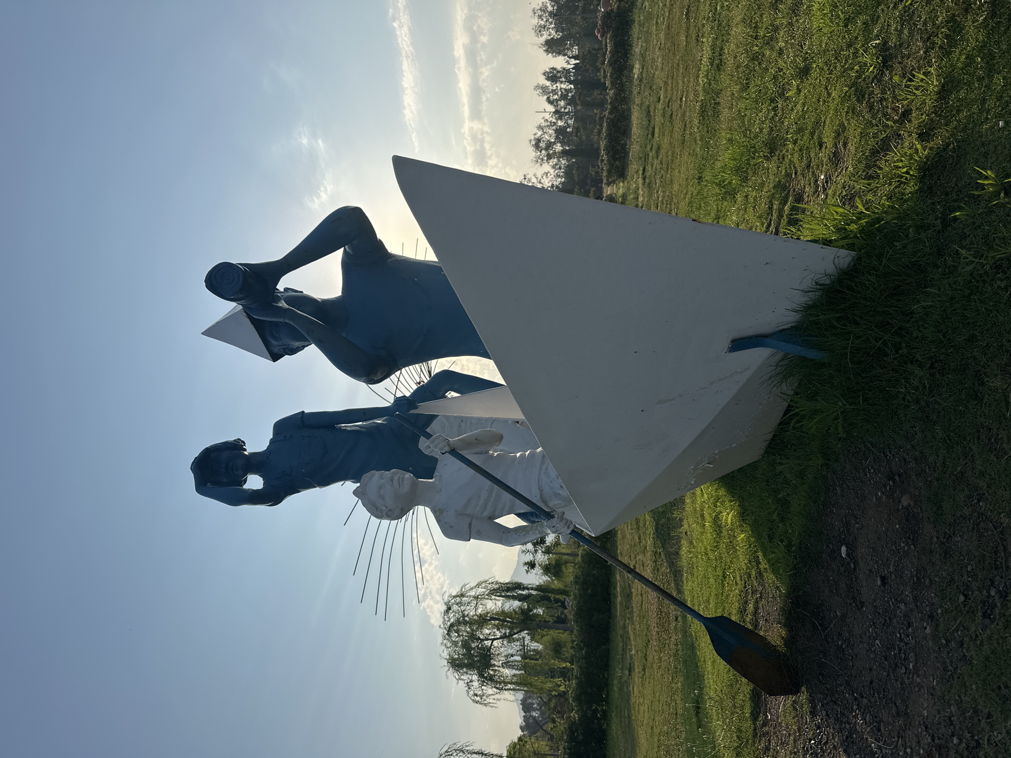 The image depicts a sculpture in Fethiye, Turkey, featuring three figures in a boat. The sculpture is set in a grassy park with trees in the background and a bright sky overhead. The figures are stylized and painted in blue and white. The front figure, painted white, is positioned as if rowing with an oar. Behind this figure, two blue figures stand. One is looking upwards with a hand shielding their eyes, and the other is holding a telescope to their eye, wearing a conical hat. The boat itself is a stylized white shape. The lighting suggests it is either sunrise or sunset, casting long shadows and illuminating the scene with warm light.