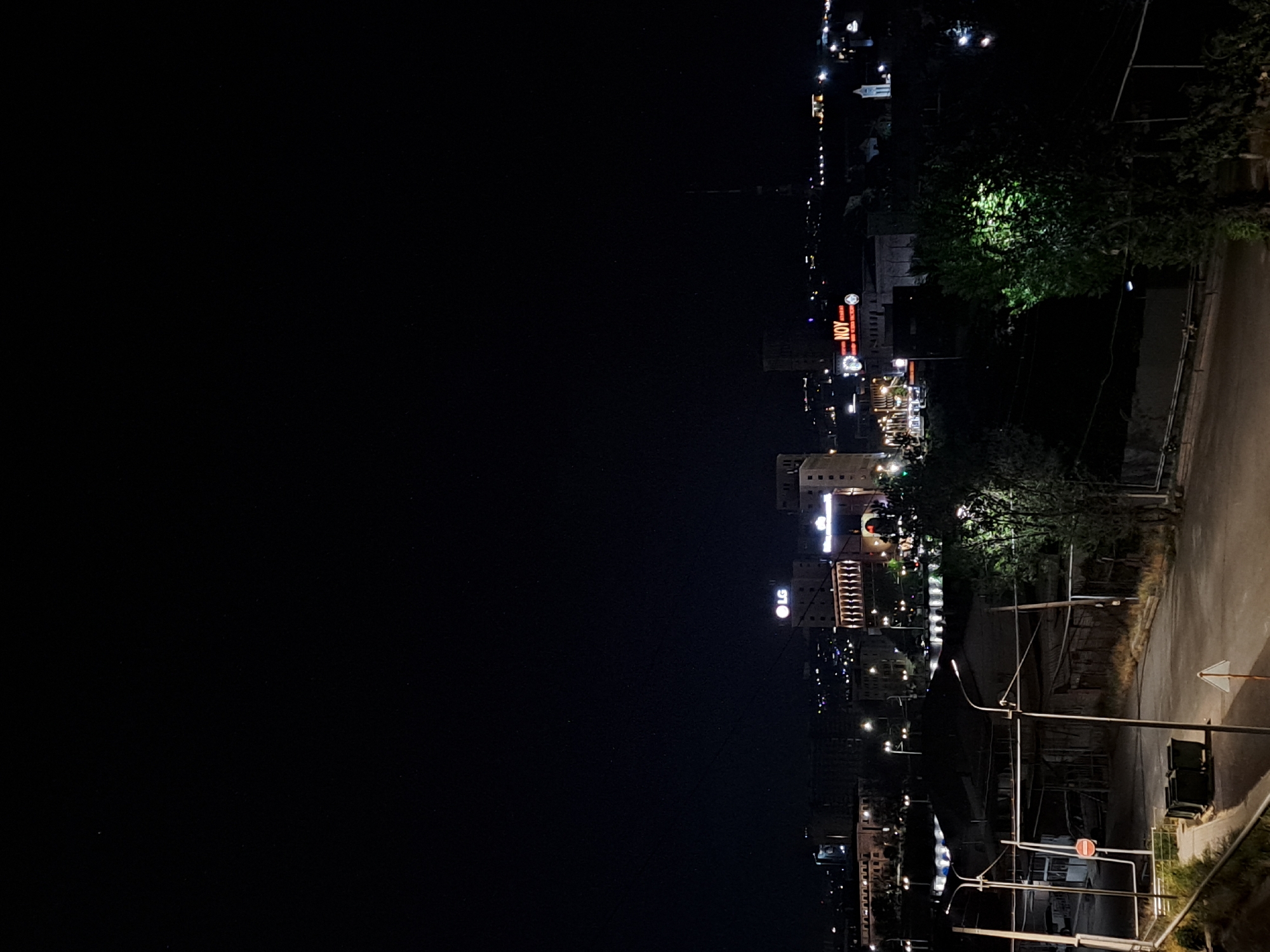 The image depicts a night cityscape in Yerevan, Armenia, featuring illuminated buildings and streetlights. Prominent in the view is the "NOV" sign atop a building, possibly indicating a restaurant or business name. Nearby, the "LG" sign is also visible on another building. The scene includes trees and a road, suggesting an urban residential or commercial area. The darkness of the night obscures most of the details, but the city lights create a distinct skyline. The image seems to be taken from a high vantage point, providing an overview of the city.
