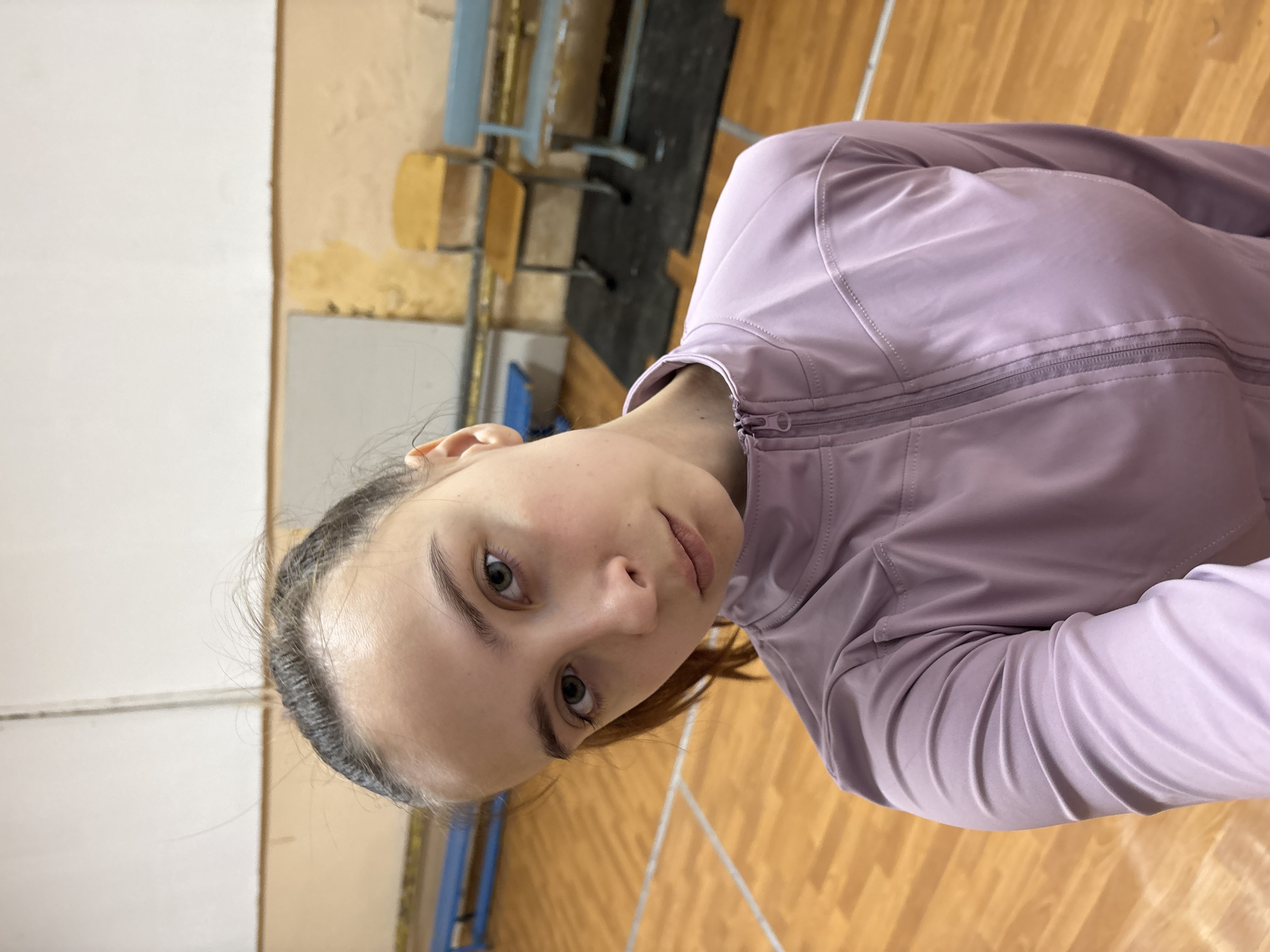 A young woman is depicted in a medium-angle shot in what appears to be a gymnasium in Chaikovsky, Russia. The background includes wood flooring with white lines and gymnasium equipment, and a light beige wall on the left. She is wearing a light purple zipped jacket. She is fair-skinned with brown hair and is facing the camera.