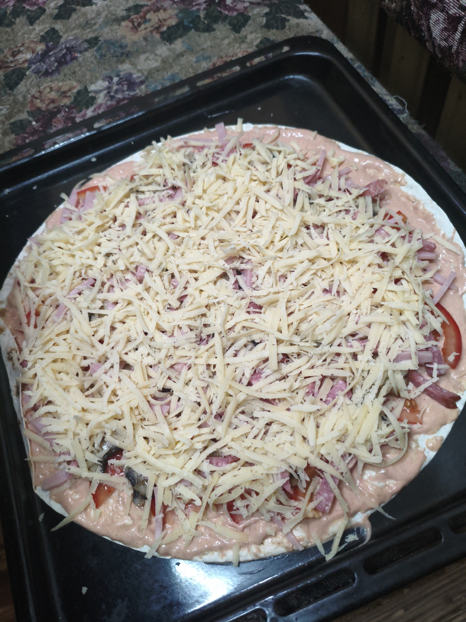 A close-up, overhead shot shows a pizza being prepared on a black baking sheet. The pizza has a thin crust, topped with a pinkish sauce, sliced tomatoes, pieces of meat, and a generous layer of shredded cheese. The baking sheet itself is placed on a patterned surface that appears to be a tablecloth. The lighting suggests an indoor setting, likely a kitchen, and there are no individuals or discernible urban elements present to indicate a specific city location.