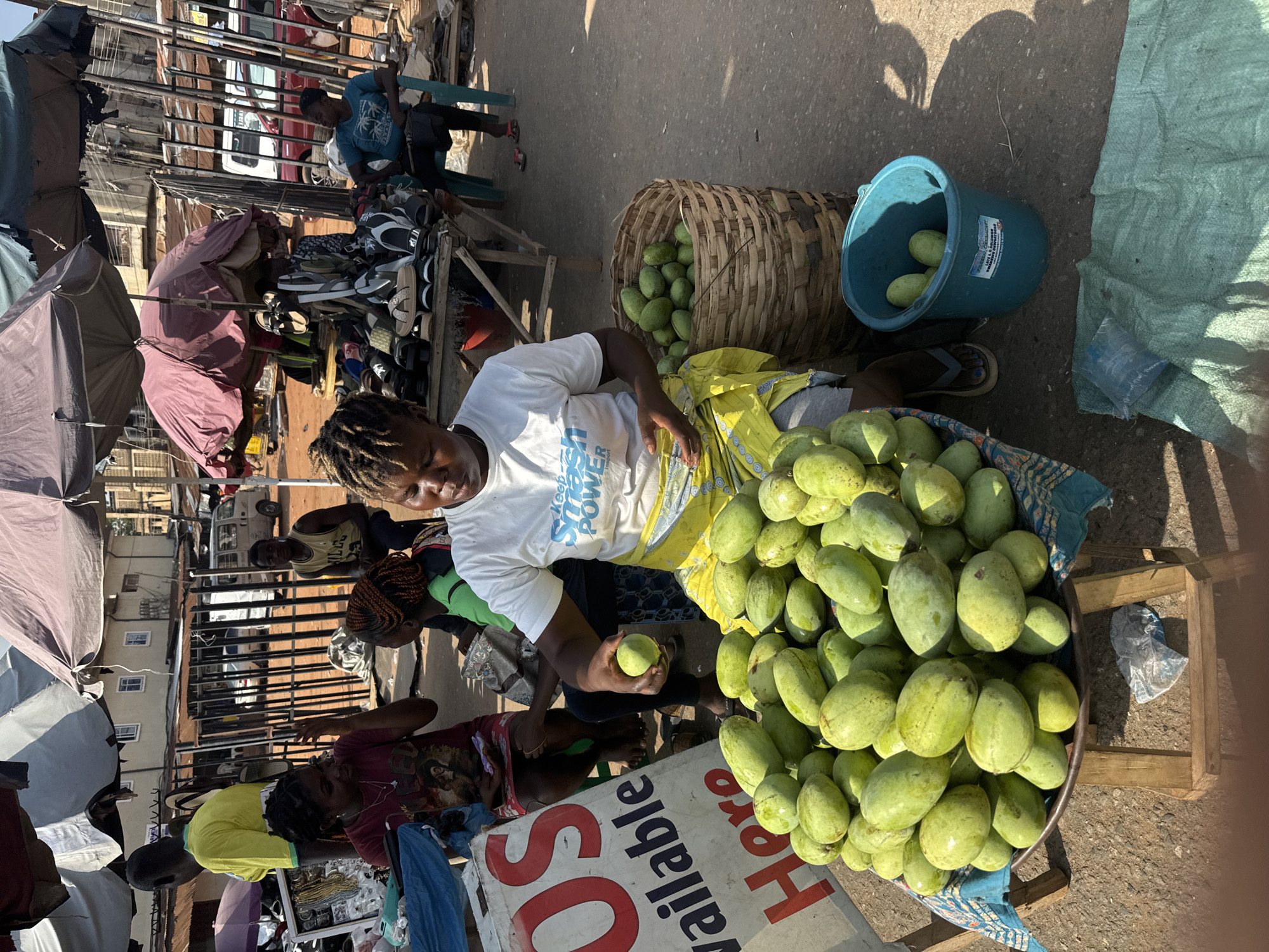 A daytime scene captures a vendor in a marketplace in Onitsha, Nigeria. The vendor, a person with braided hair, is wearing a white t-shirt. They are seated beside a large basin filled with green mangoes. The vendor holds one mango in their hand. A woven basket and a blue bucket, both containing more mangoes, are nearby. A sign advertising "SOS Valuable Here" is visible. In the background, other vendors are present, selling items like footwear under umbrellas. The setting appears to be a bustling open-air market within the urban environment.