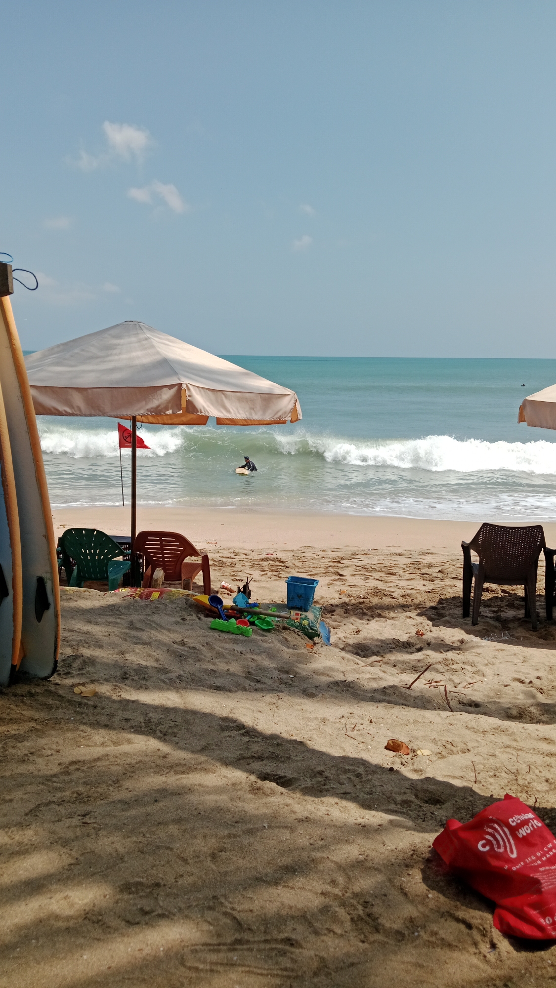 This is a medium shot of Kuta Beach in Bali, Indonesia, on a clear day. In the foreground, sand fills the bottom half of the image, with shadows cast by beach umbrellas. Scattered on the sand are various children's toys, including buckets, shovels, and colorful plastic items. Two empty plastic chairs and one empty chair are visible near a large beach umbrella. Two surfboards stand upright on the left side of the frame.
In the middle ground, the sandy beach meets the ocean. Waves are breaking, and a person is seen in the water, appearing to be bodyboarding or surfing. A red flag is planted on a pole near the umbrella.
The background is dominated by the turquoise ocean and a clear blue sky with a few small white clouds. The horizon line where the ocean meets the sky is clearly visible. The overall scene depicts a typical day at a popular Indonesian beach, with activities and objects associated with leisure and water sports.