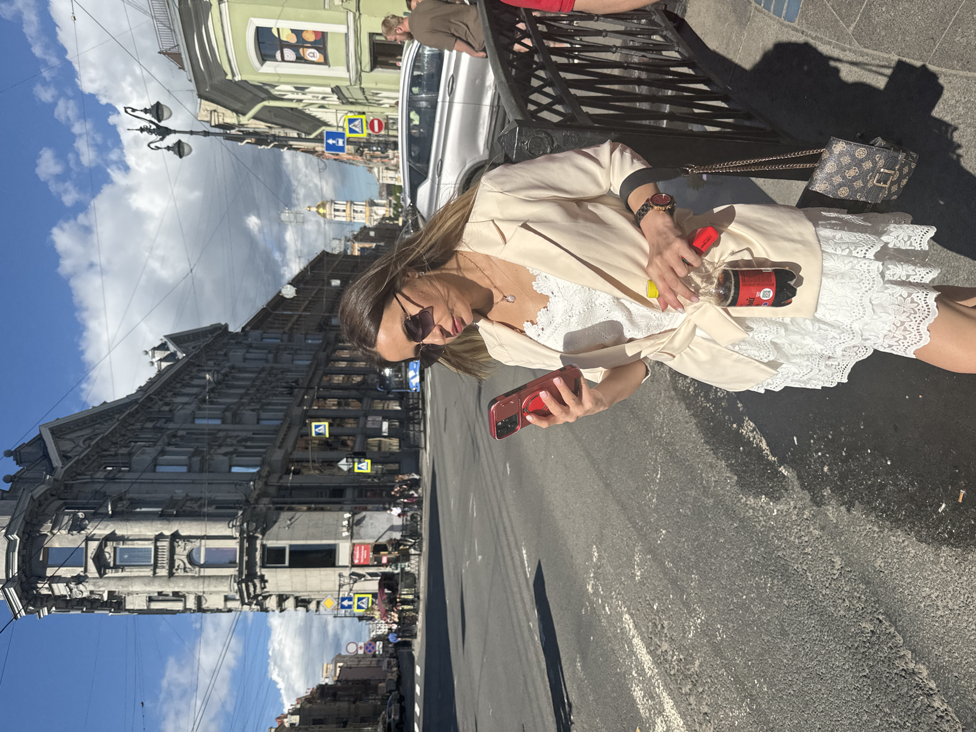 A woman in a cream-colored blazer and white lace dress stands on a city street in Saint Petersburg, Russia, looking at her red smartphone. She holds a bottle of dark soda and a red object, possibly a vape or lipstick. A designer handbag hangs from her shoulder. In the background, a silver car, pedestrians, and a distinctive building with a golden dome are visible under a cloudy blue sky.