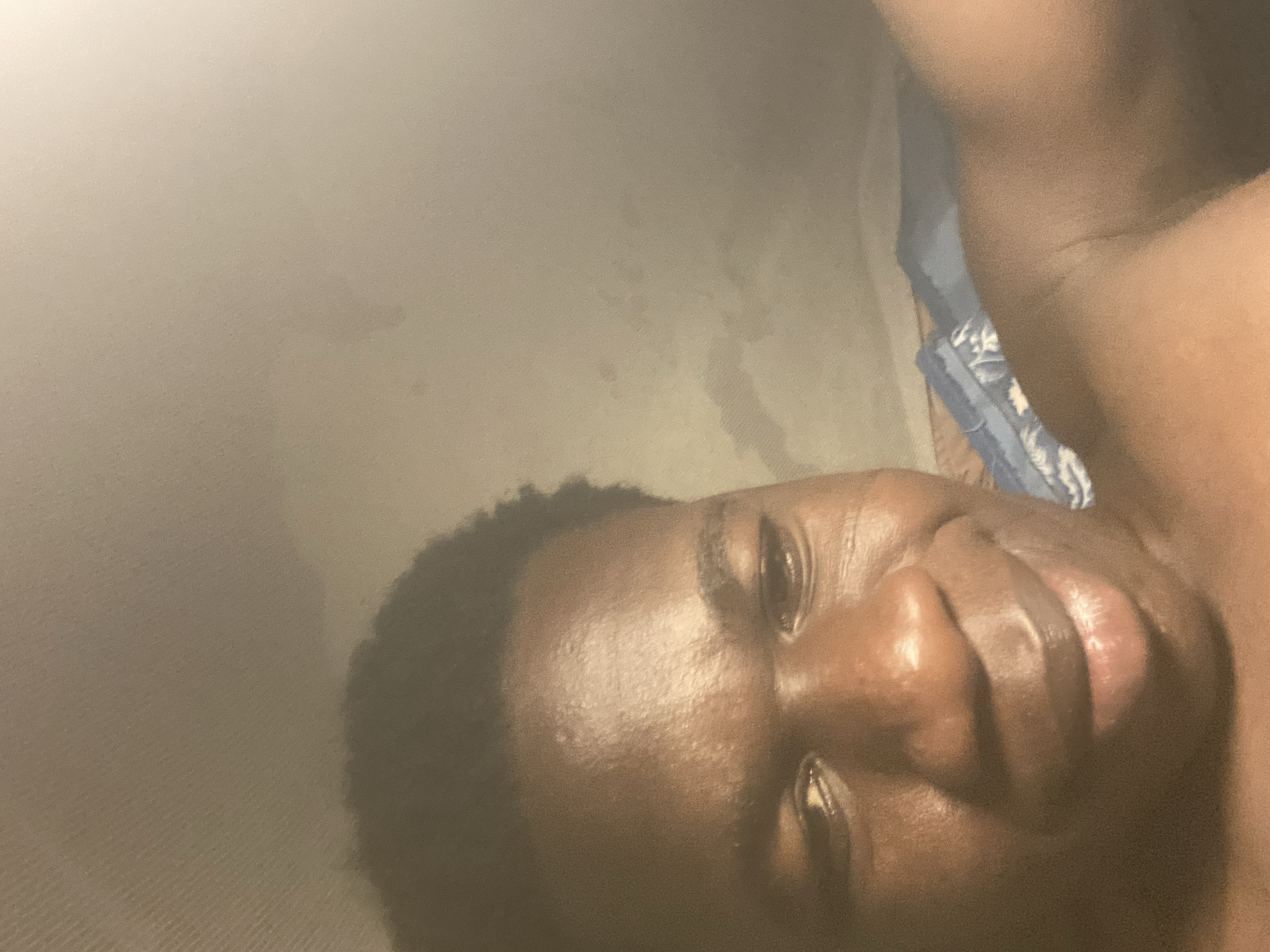 The image shows a close-up of a dark-skinned person's face, likely male, lying on a surface. The face is angled towards the camera. Part of a bed or bedding is visible in the top right corner. The image has a slight yellow tint. There is no information to indicate the photo was taken in Mararaba, Nigeria.