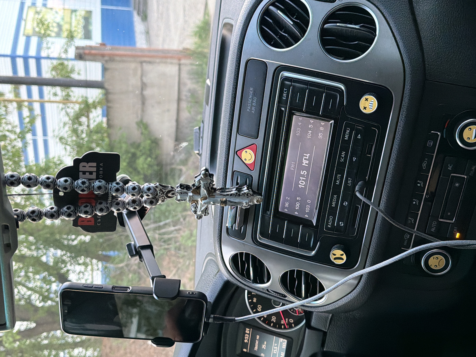 Here is a detailed description of the image:
**Setting:** The image appears to be taken from inside a vehicle, specifically the driver's perspective of the dashboard and front windshield. The car seems to be parked.
**Environment:** Through the windshield, one can observe an outdoor scene with buildings and greenery. There is a structure with blue and white stripes, possibly an awning or part of a building facade. There's also a building with gray exterior and some trees or bushes.
**Objects in the Car:**
* **Dashboard:** The car's dashboard is visible, featuring air vents, a radio/media system, climate control buttons, and instrument panel.
* **Radio/Media System:** The radio display shows the frequency 101.5 MHz and some other frequency presets. There are buttons for "RADIO," "MEDIA," "MUTE," "SCAN," "MENU," and "LIST."
* **Climate Control:** The climate control panel has buttons for "AUTO," "AC," "OFF," and "DUAL."
* **Instrument Panel:** The speedometer, other gauges, and digital display showing "9:53 01" are visible.
* **Car Accessories:**
* A phone mount holding a smartphone.
* A beaded rosary with a cross hanging from the rearview mirror.
* A car freshener/air freshener accessory is also visible.
**Other details:**
* There are "smiley face" stickers on some of the buttons.
* A cable is plugged into the smartphone.
* A "PASSENGER AIR BAG OFF" indicator is near the right side of the dashboard.