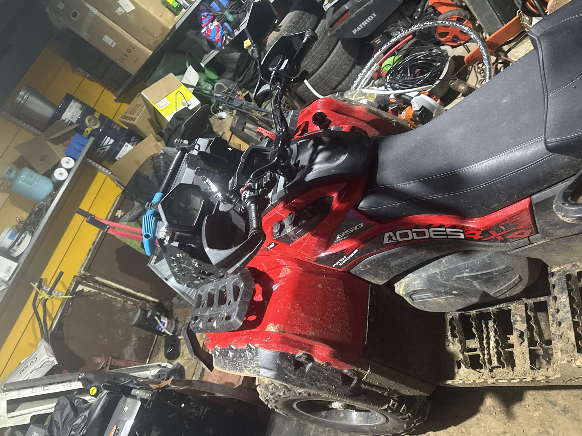 The image shows a red and black ATV parked in what appears to be a cluttered garage or workshop. The ATV has mud on its tires and body, suggesting it has been used recently. Behind the ATV, shelves are visible, filled with various items including boxes, tools, and what looks like a gas cylinder. The walls of the structure are painted yellow. The overall impression is of a working space that is somewhat disorganized. The scene is devoid of any people.