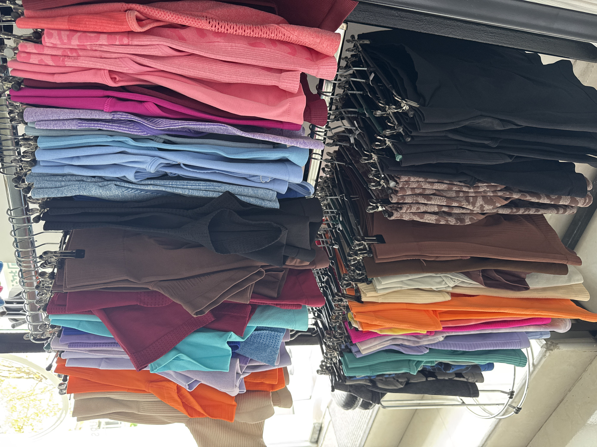 This image shows a retail display of women's activewear, specifically a rack of colorful athletic shorts and tops, in a store located in Batumi, Georgia. The clothing is organized by color, ranging from neutral tones like beige and brown to vibrant shades of orange, turquoise, purple, pink, and red. The items are hung on metal hangers on a double-tiered display rack. The background suggests an indoor retail environment with a window providing natural light and glimpses of foliage outside. The scene is static, with no people visible in the frame.